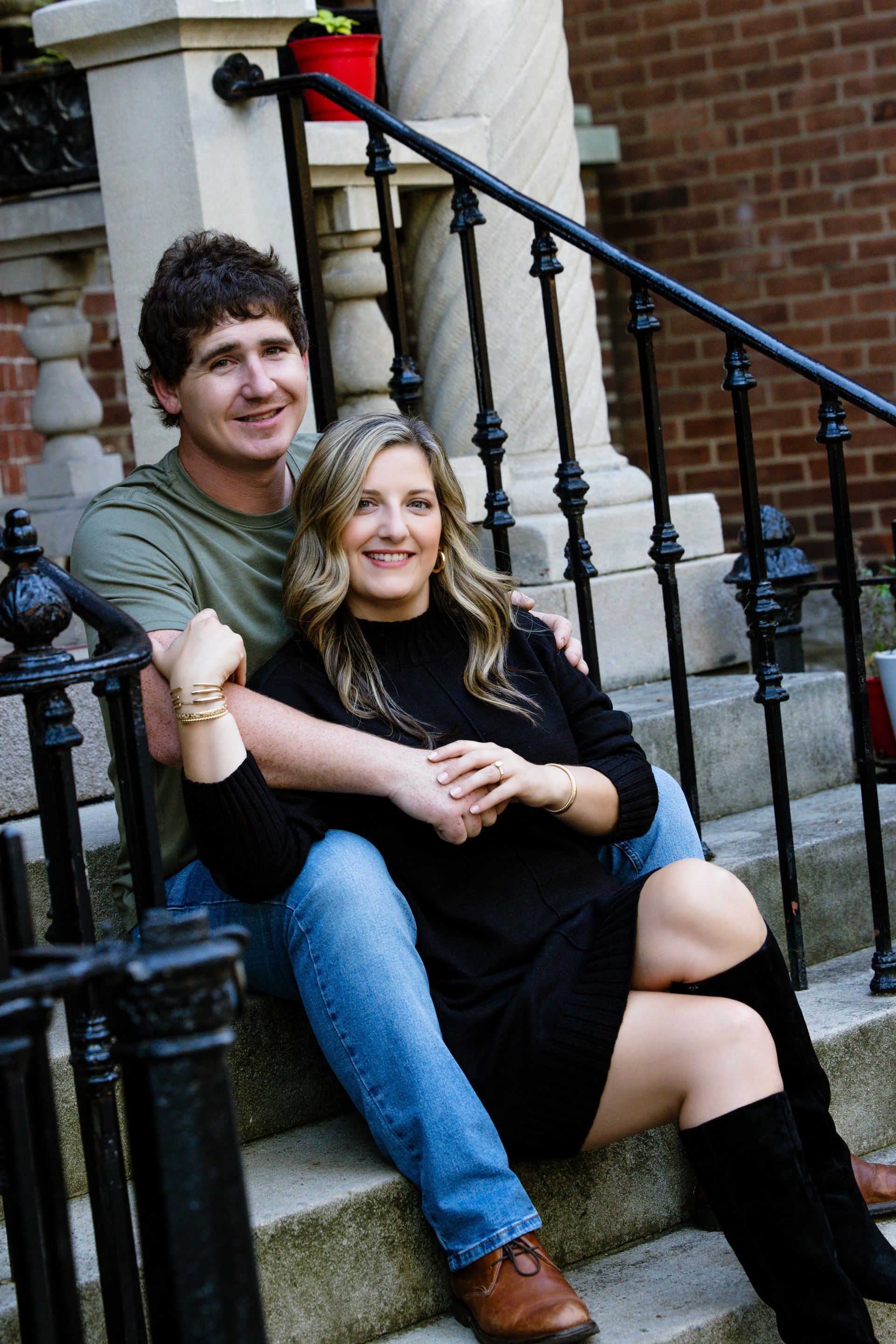Couple sitting on stone steps outdoors, smiling in front of a brick building and black railing