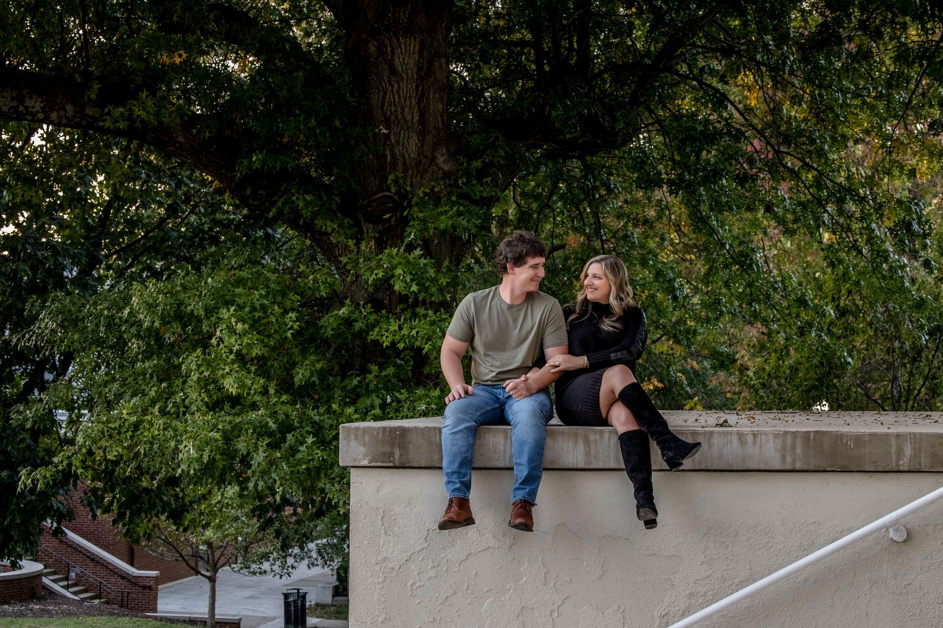 Two people sitting on a concrete ledge outdoors, talking in front of dense green trees.