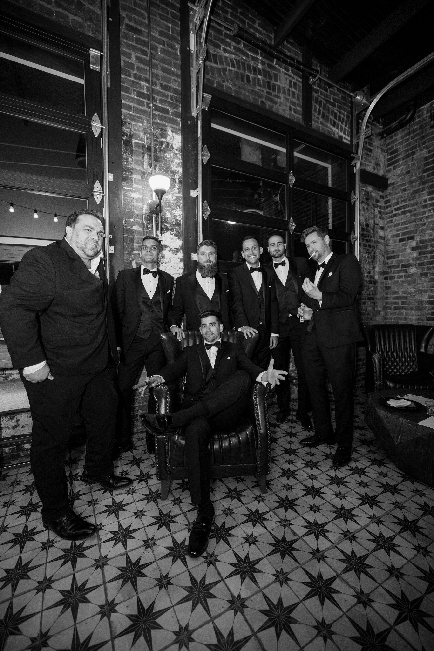 Group of people in formal suits posing in a brick-lined interior with patterned tile floor, black-and-white photo