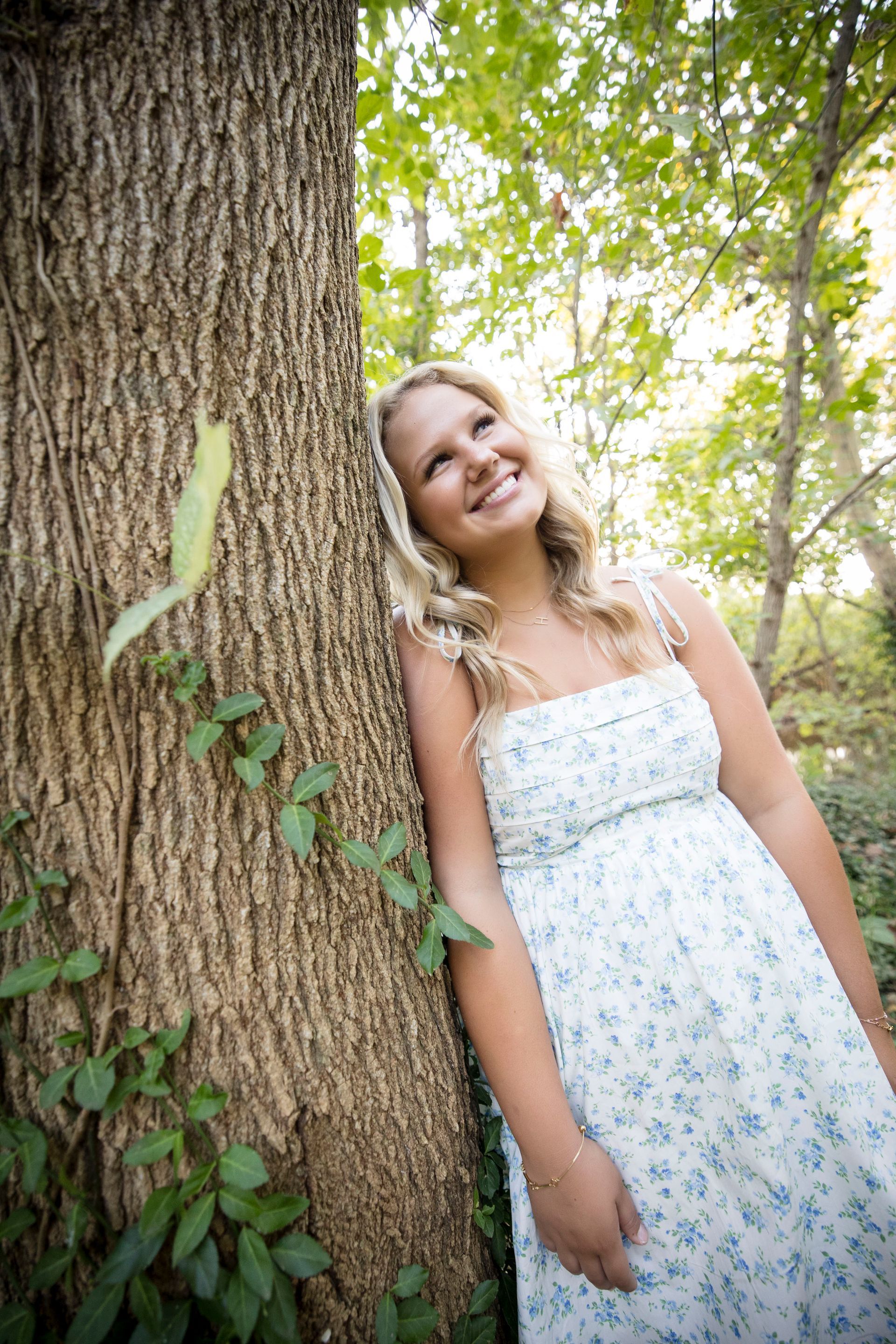 Woman in a white floral dress leaning against a tree in a sunlit forest, smiling upward