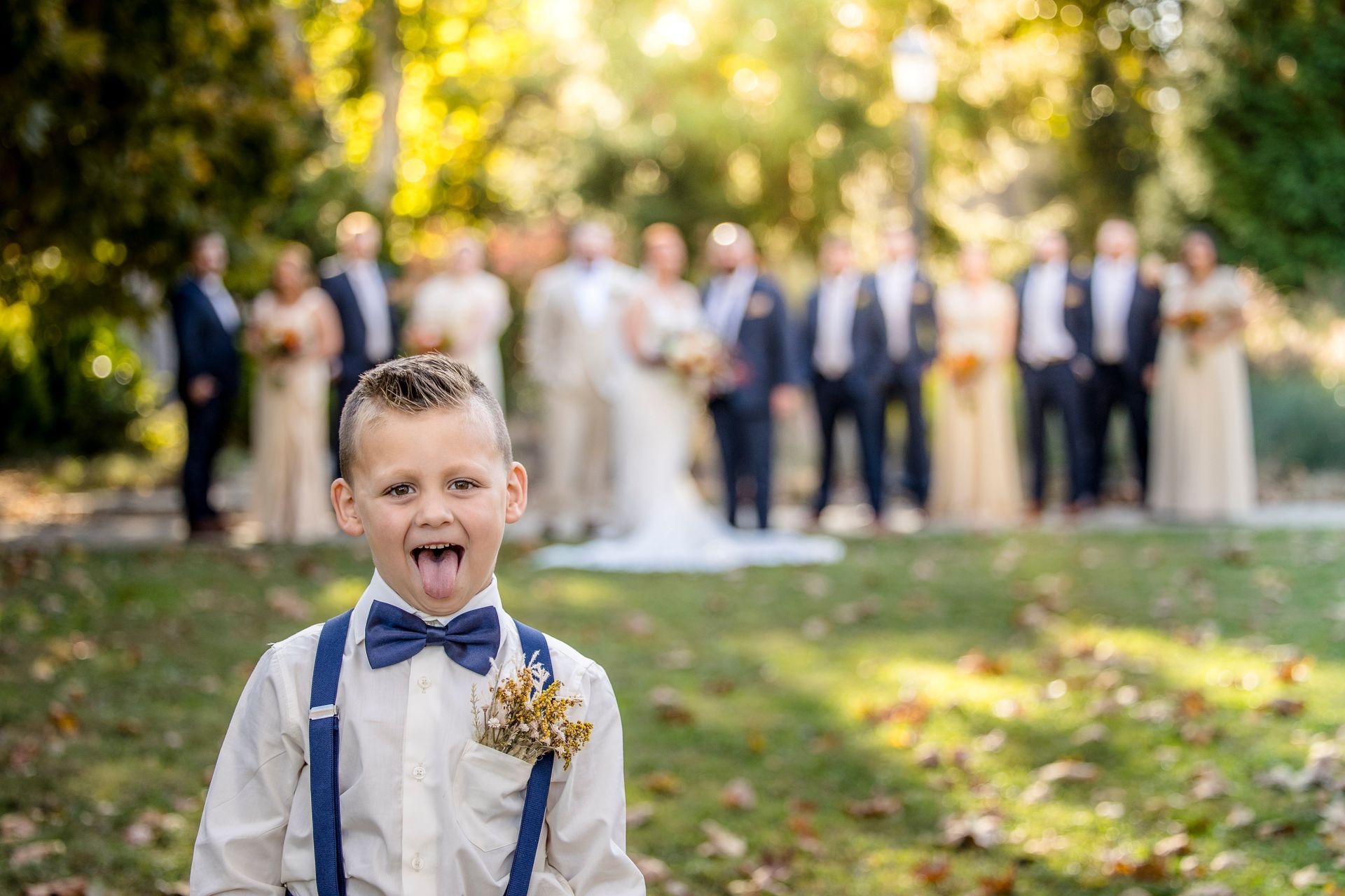 Young boy in suspenders and bow tie sticks out his tongue at a wedding party outdoors.