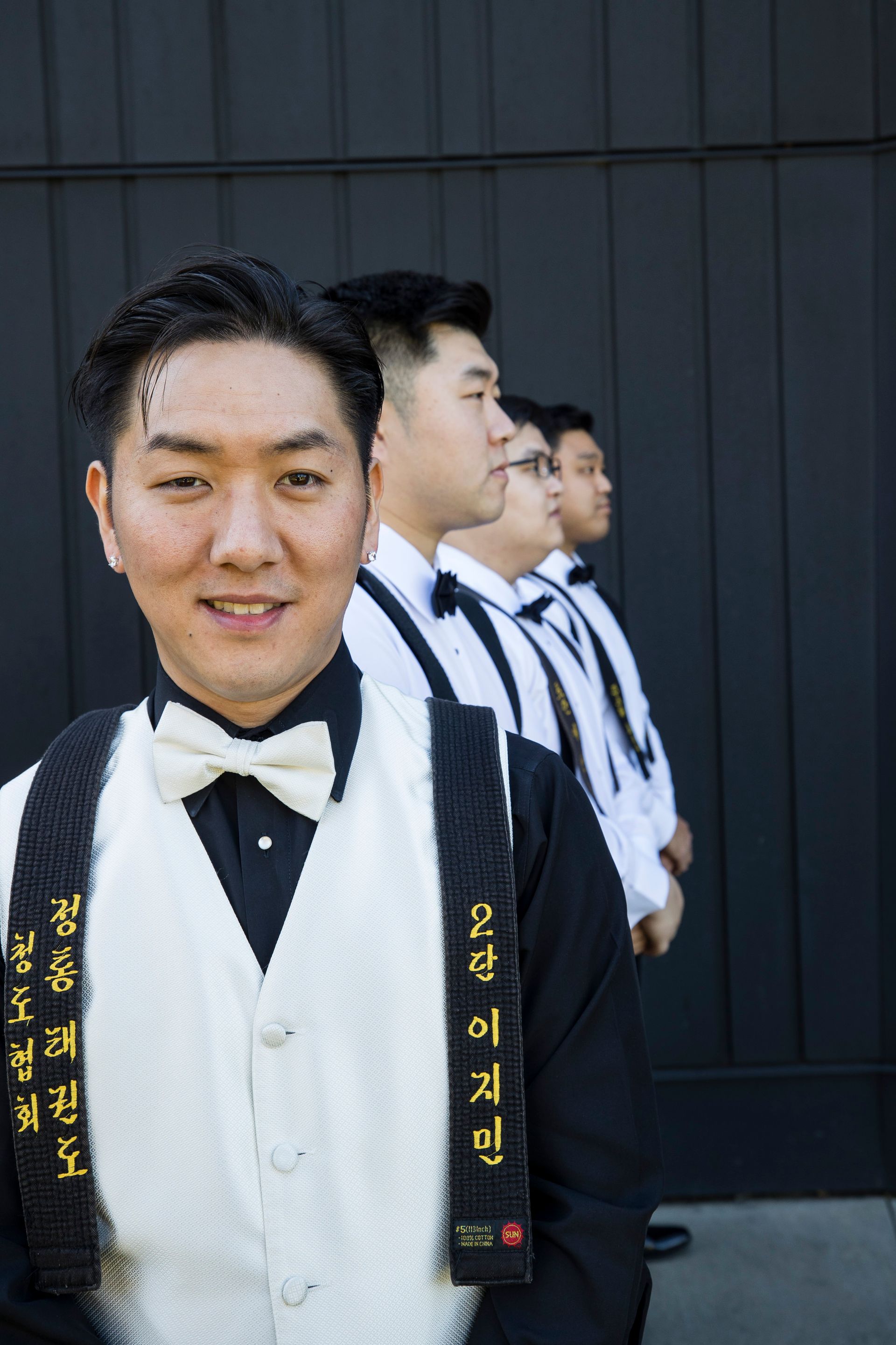 Four men in formal black-and-white attire stand in a line; the front man faces the camera smiling.