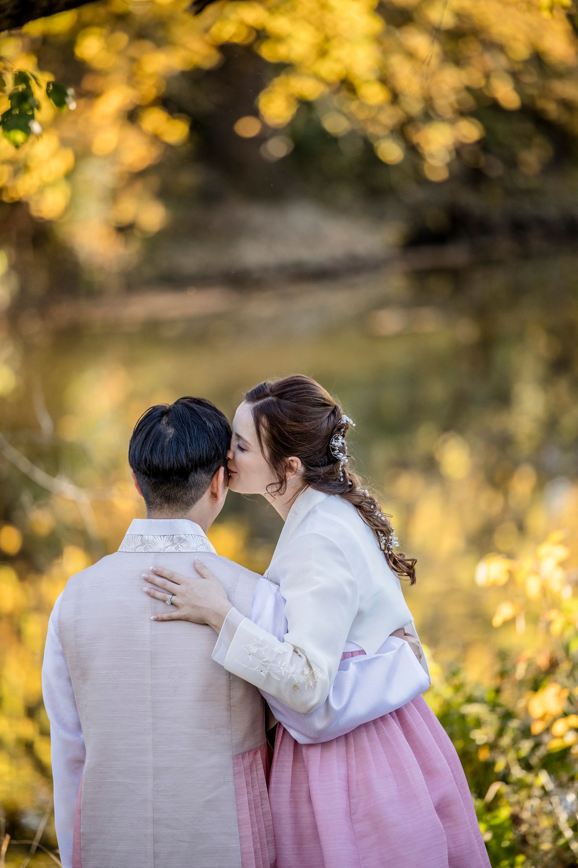 Couple in white and pink embraces by a river with golden autumn trees blurred behind them