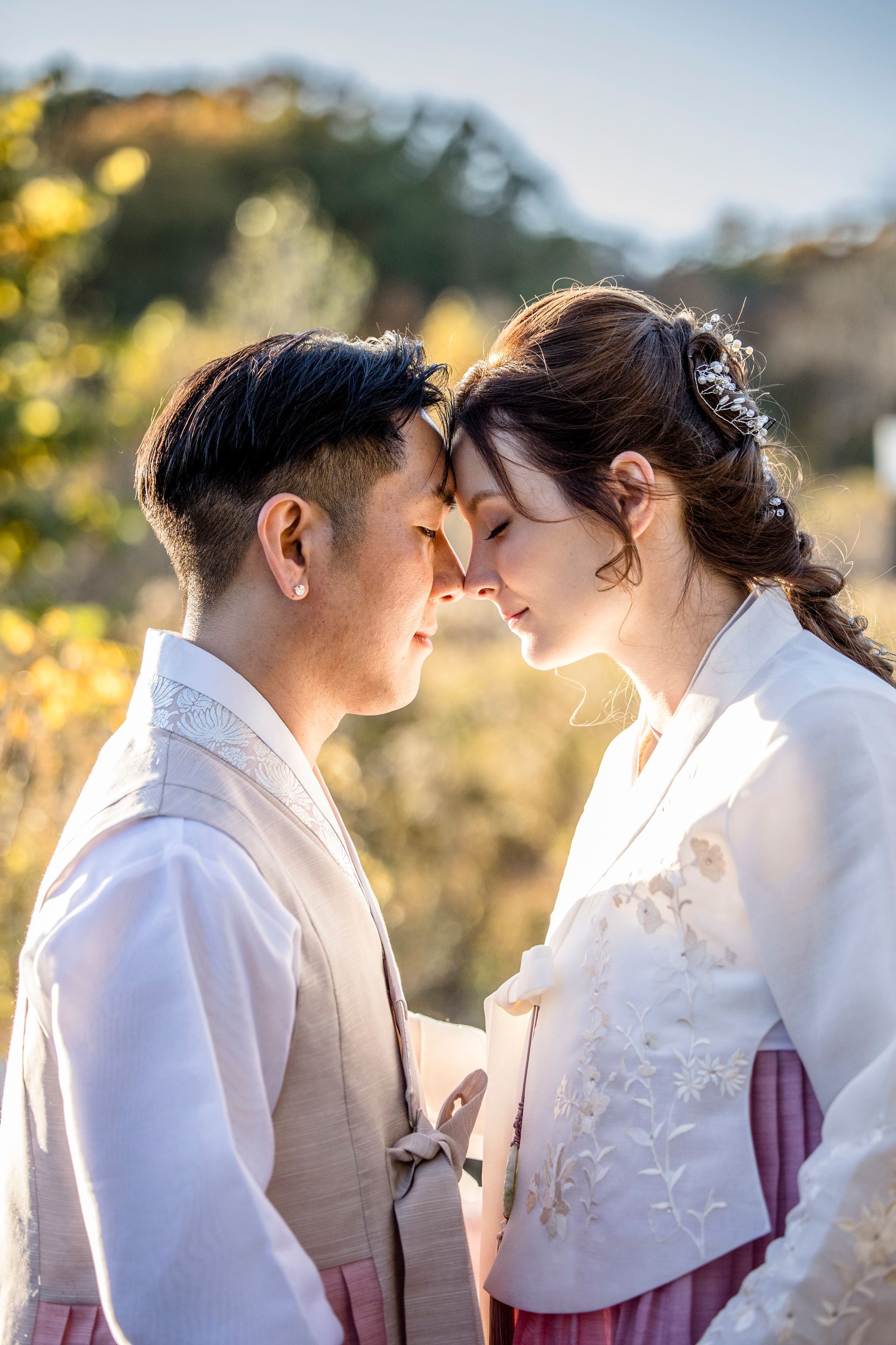 Couple in white traditional attire touching foreheads outdoors in soft sunlight