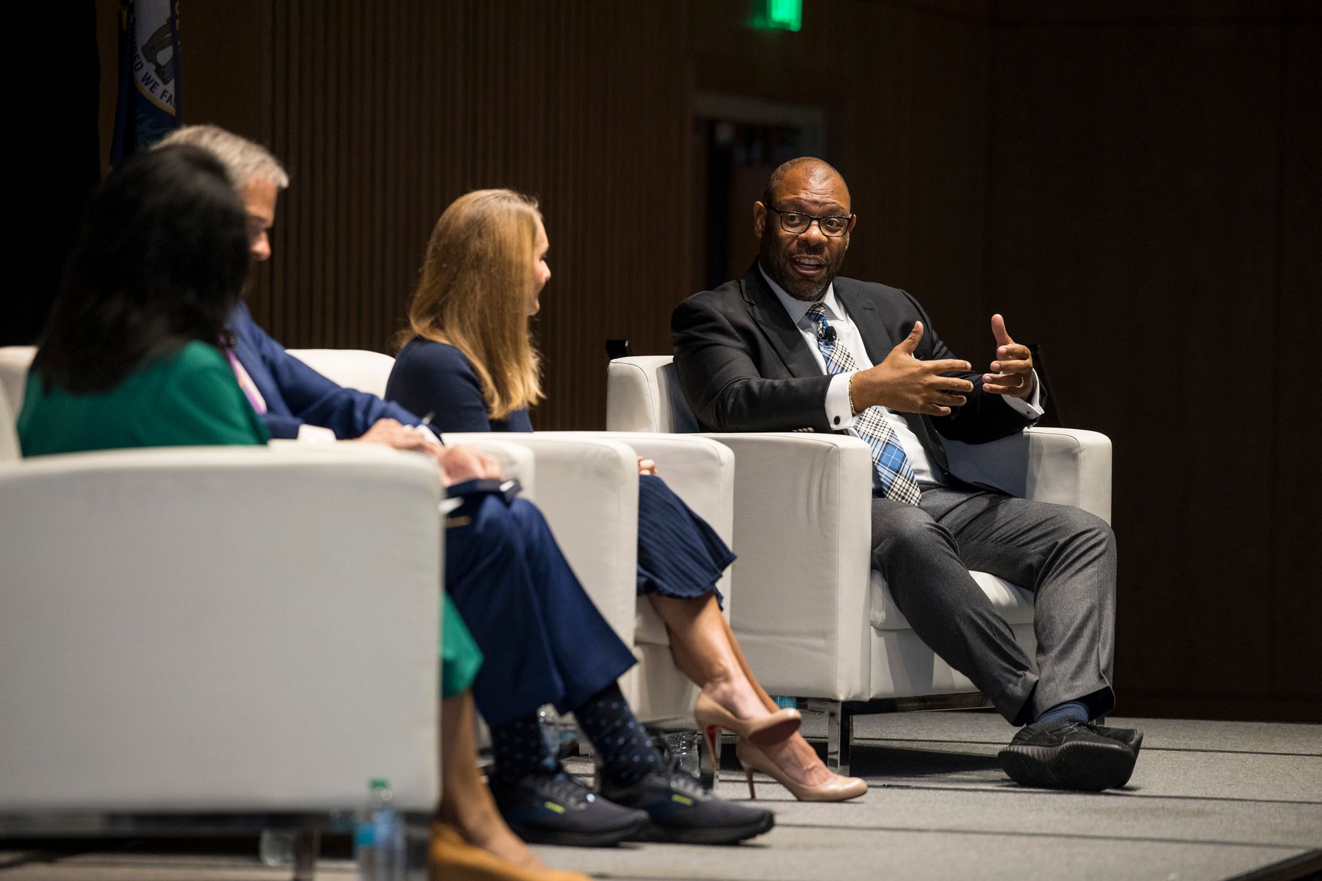 Panel discussion on stage with four seated speakers in armchairs, one gesturing while speaking