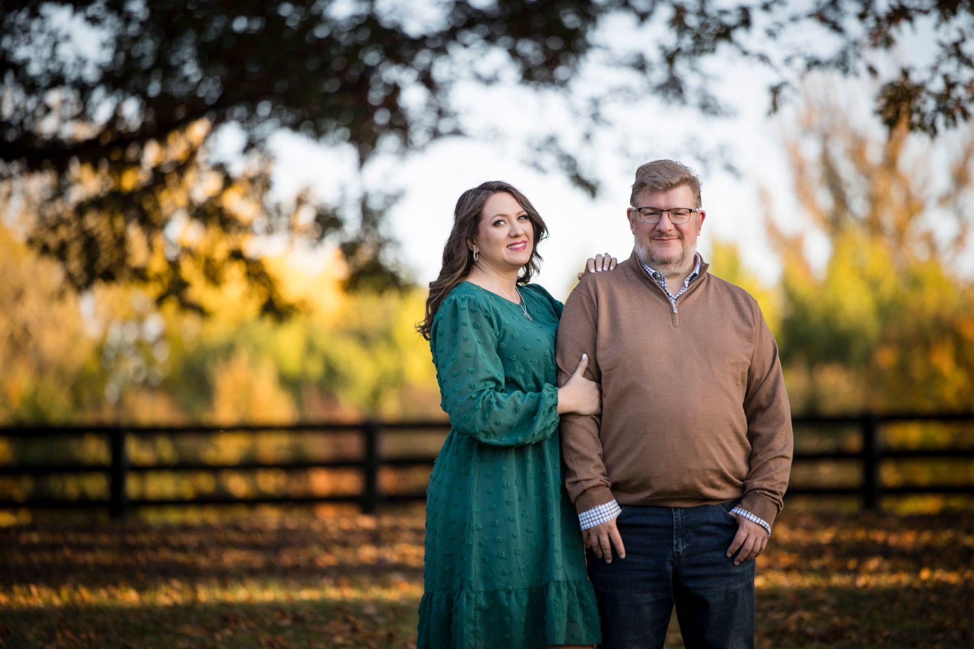 Couple standing outdoors by a wooden fence, woman in green dress and man in brown sweater, both smiling