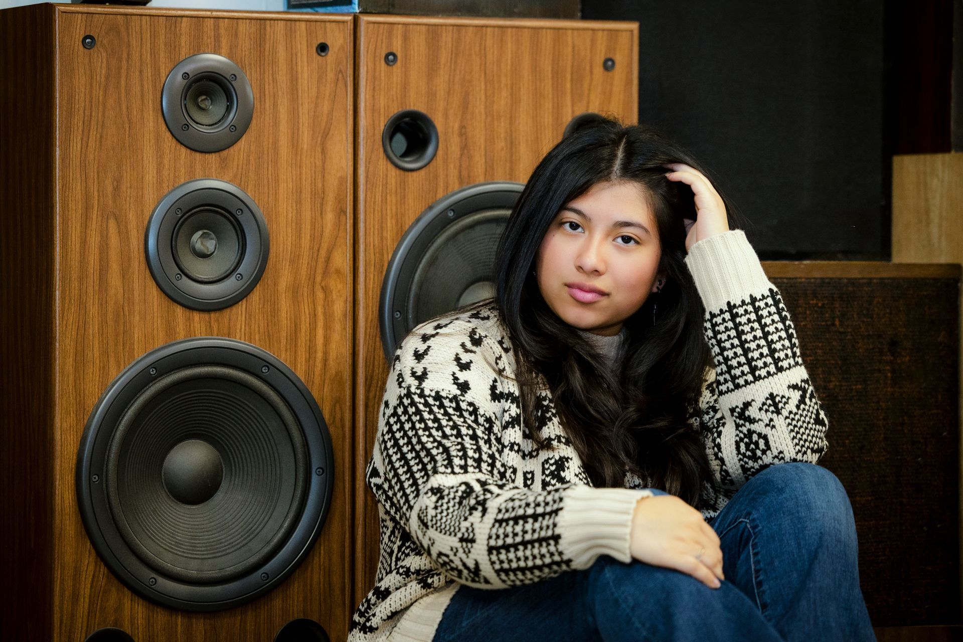 Woman sitting beside large wooden speakers in an indoor setting, wearing a patterned sweater and jeans.