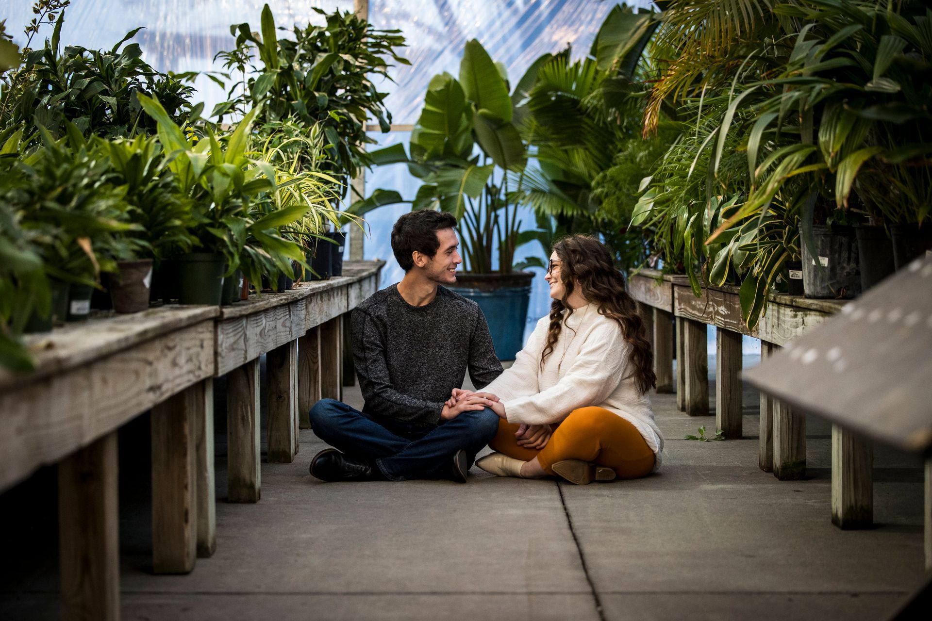 Couple sitting cross-legged on a greenhouse walkway, holding hands among lush potted plants.