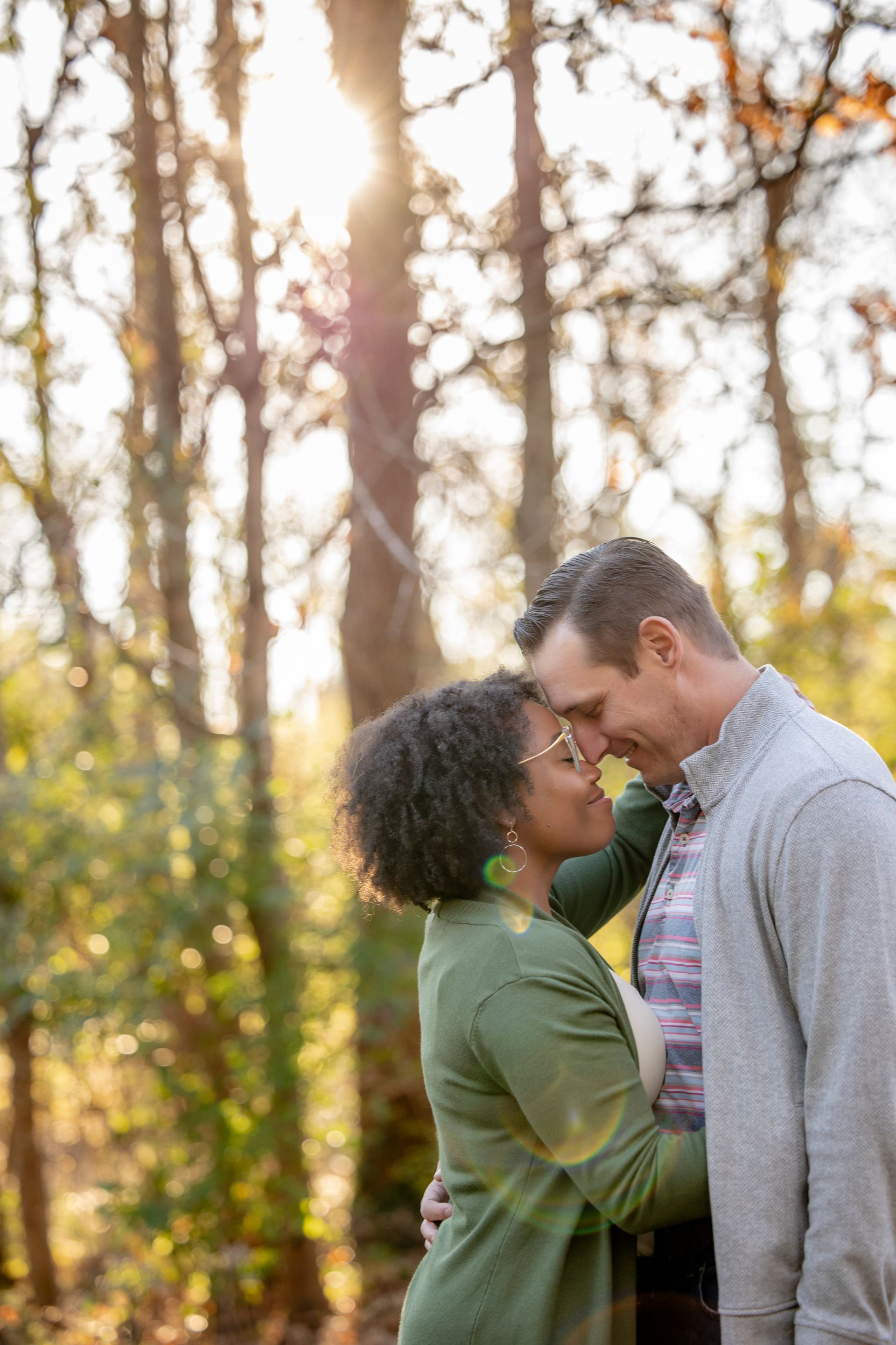 Couple standing close together in a sunlit forest, touching foreheads and smiling.