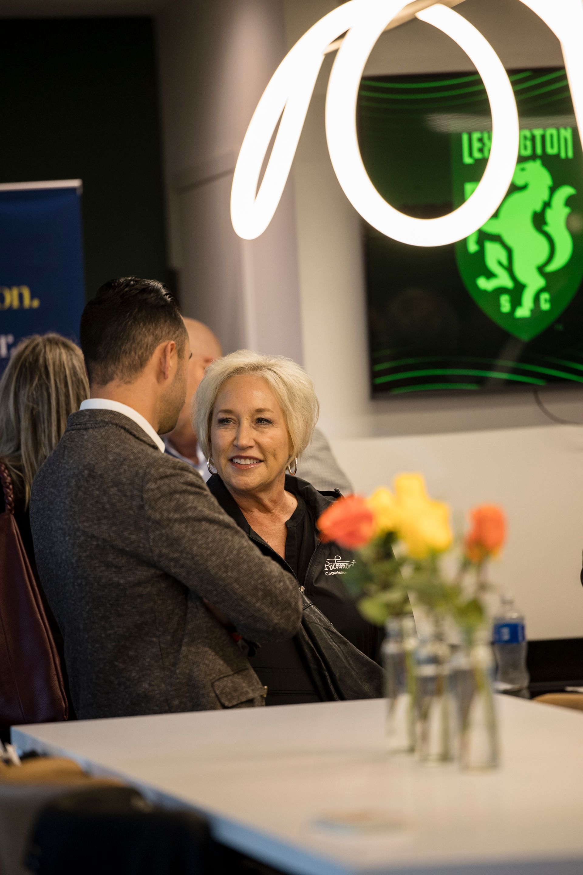 Two people conversing at an indoor event with flowers in the foreground and a glowing green sign behind them