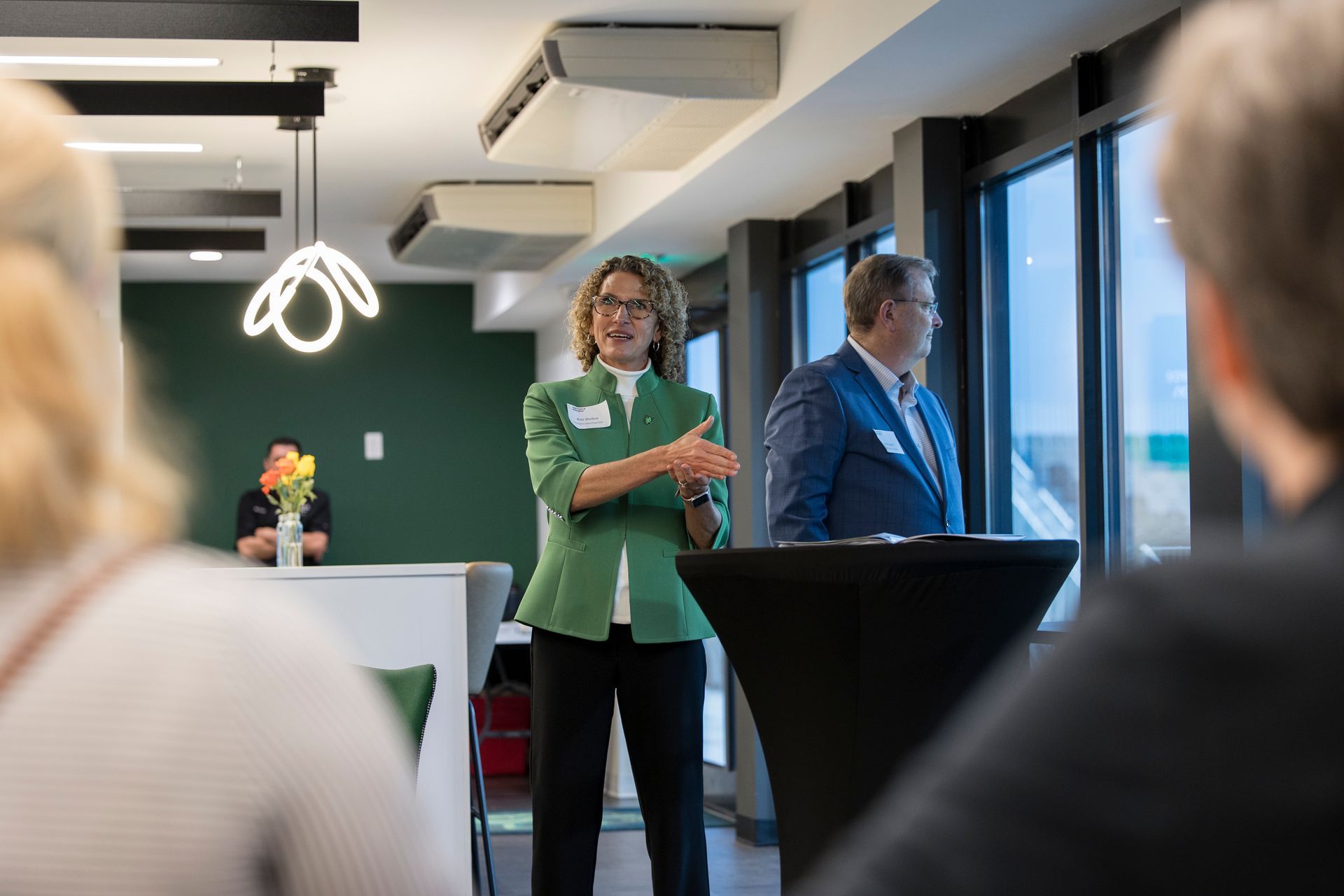 Woman in green speaking at a podium during a presentation, with a man standing beside her and audience in foreground