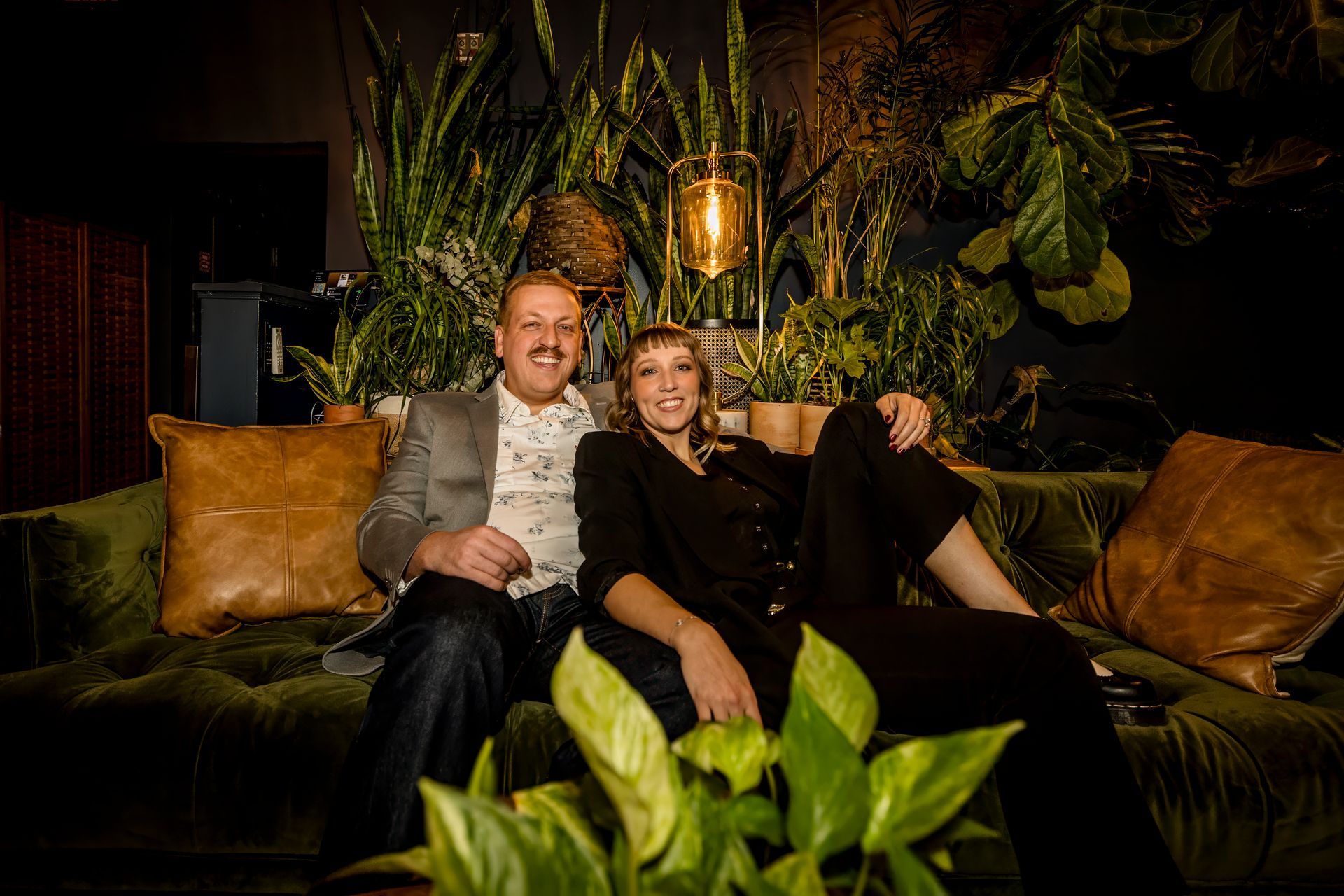 Two people sitting on a lush green couch with tan pillows in a dim, plant-filled lounge.