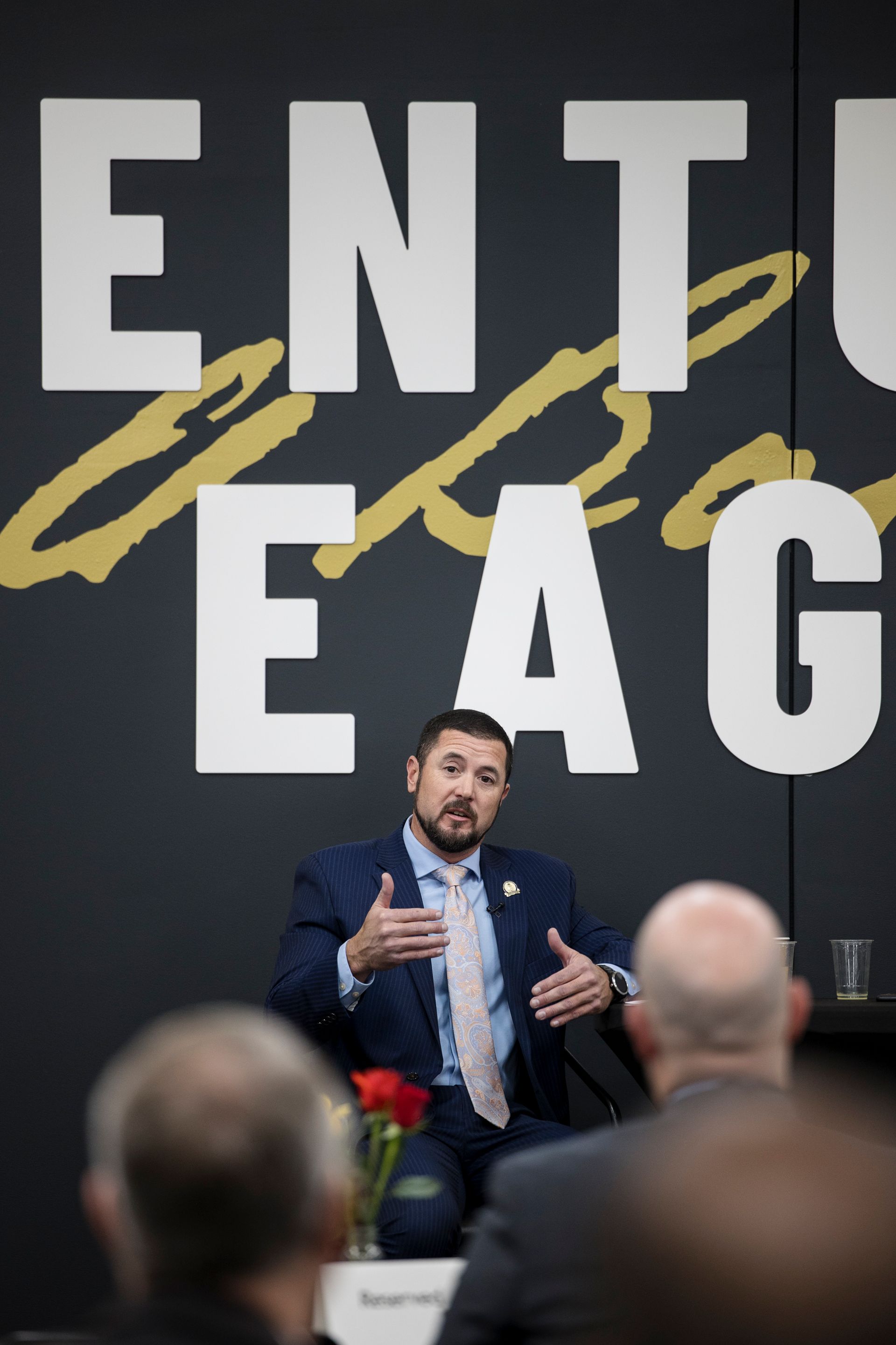 Man speaking on stage before a black backdrop with large white letters and gold accents