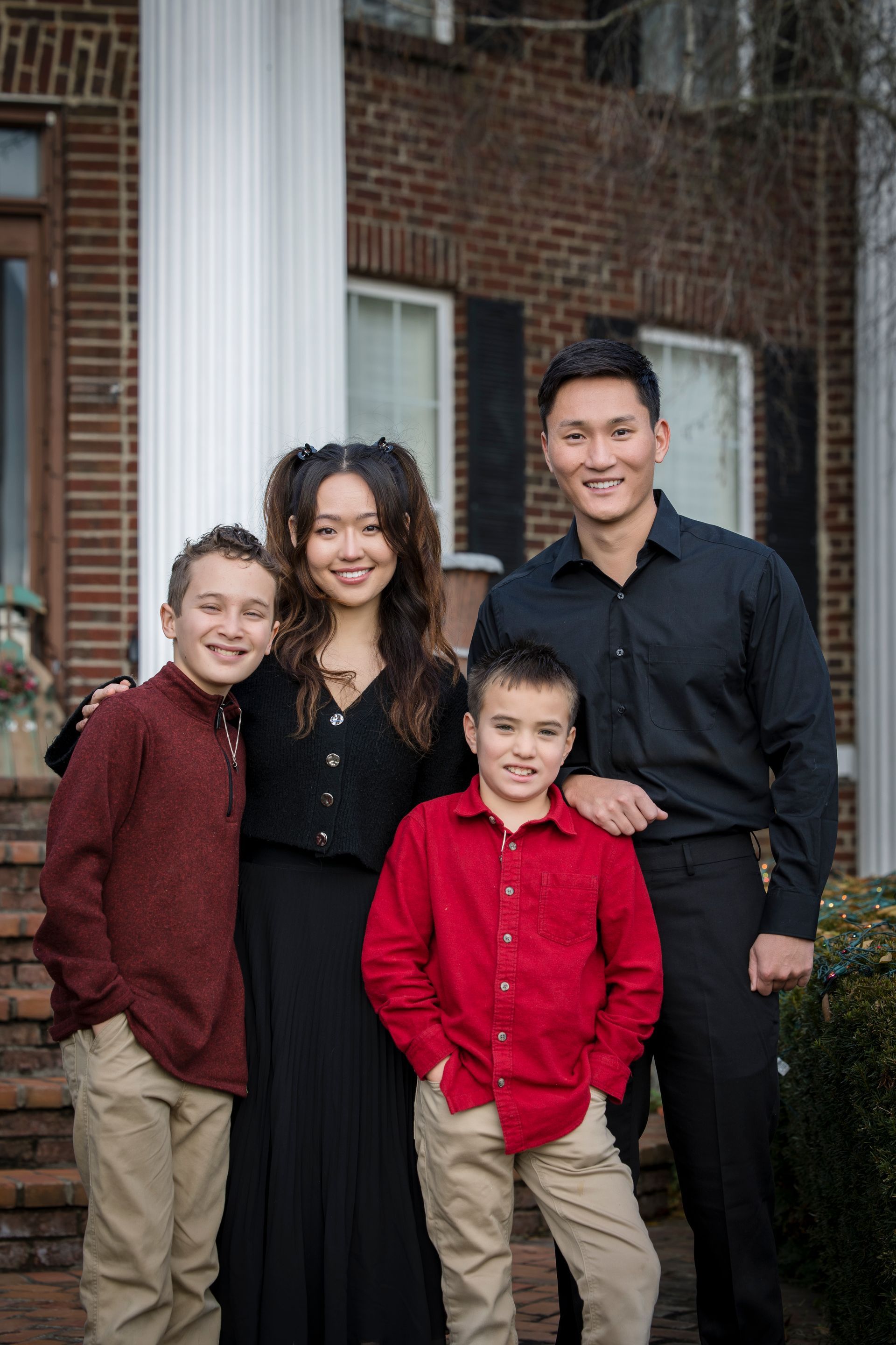 Four people posing together outside a brick house, dressed in black, red, and tan.