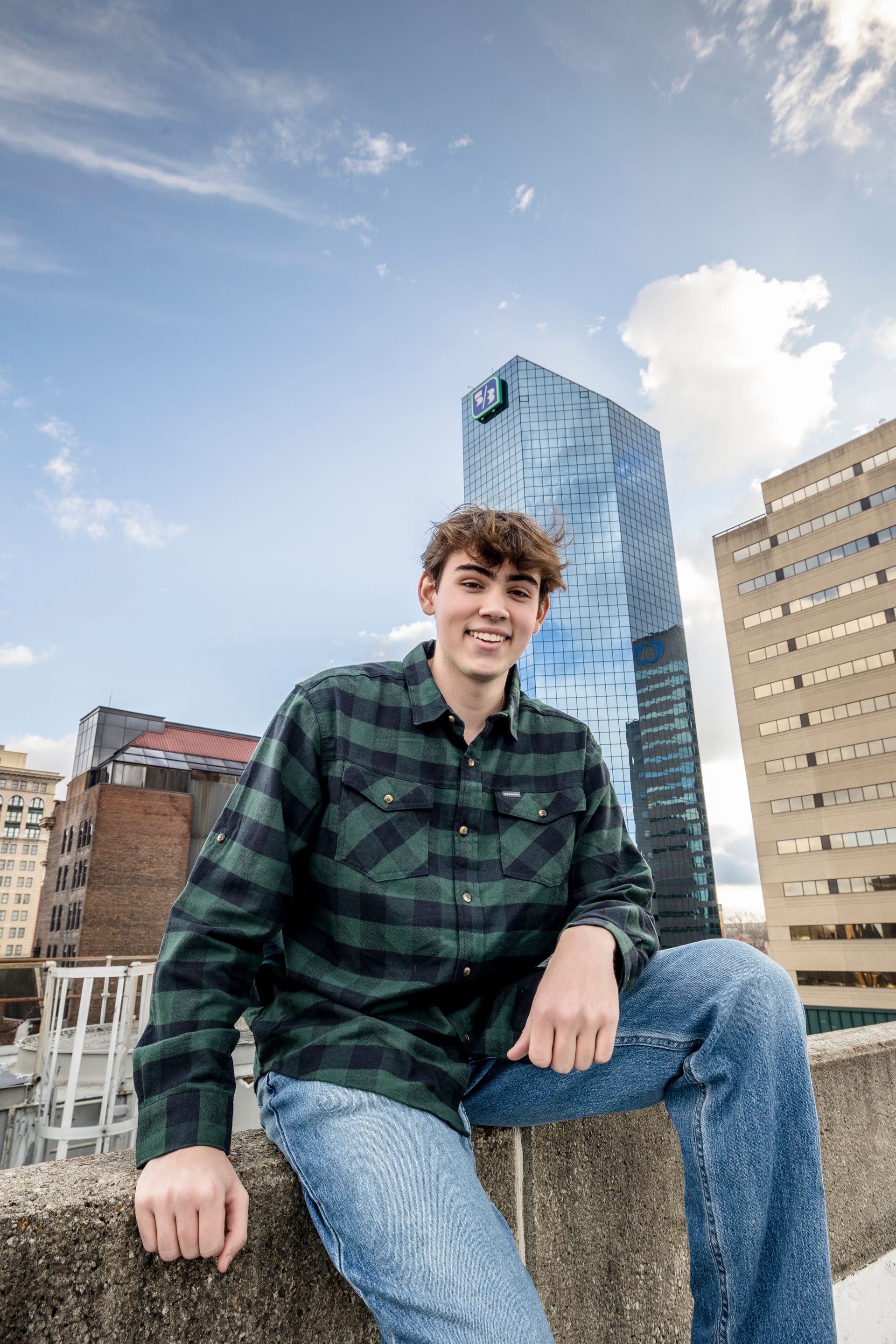 Smiling person in a green plaid shirt and jeans sitting on a ledge with city buildings and blue sky behind
