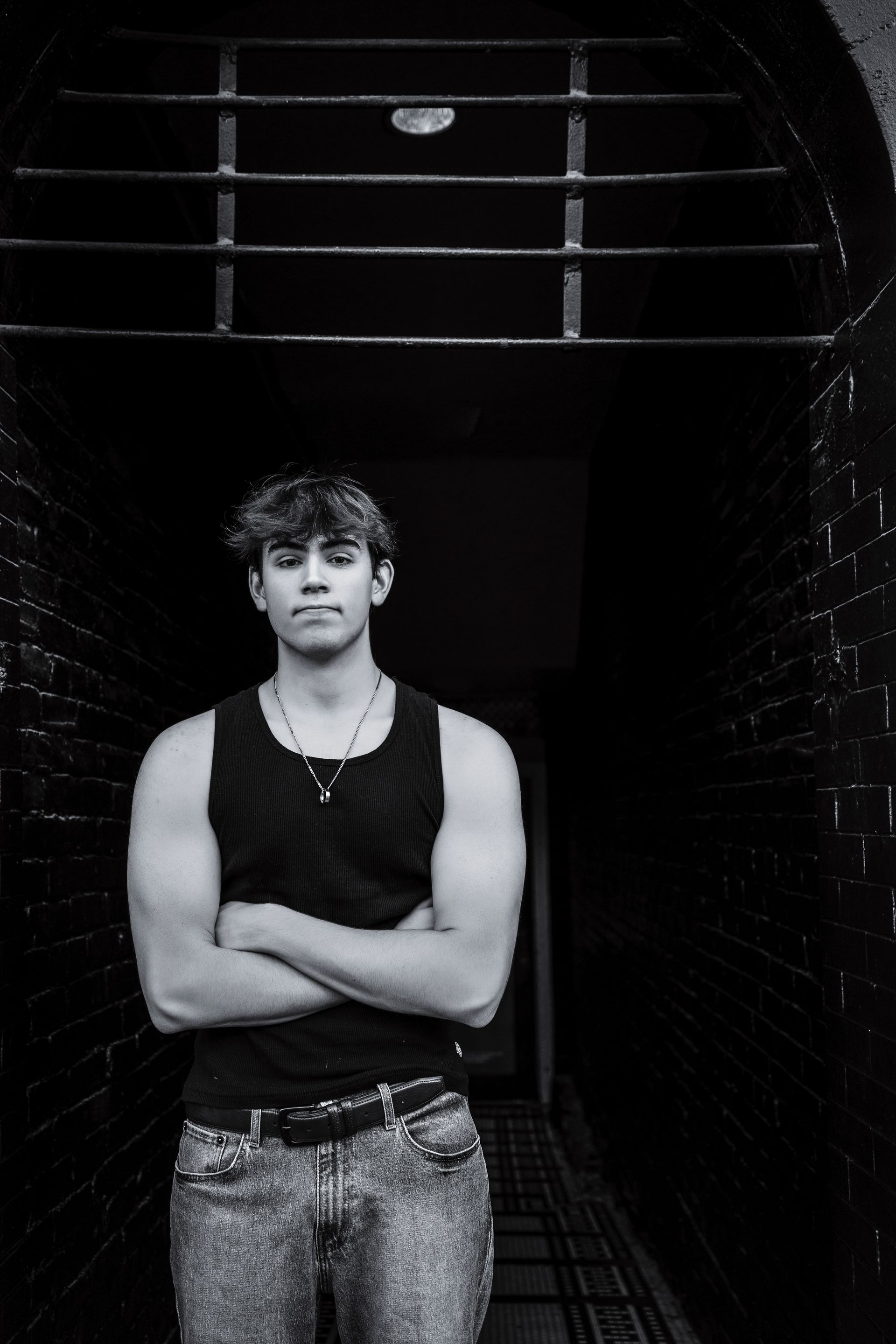 Person standing in a dark corridor with arms crossed, wearing a black tank top and jeans.