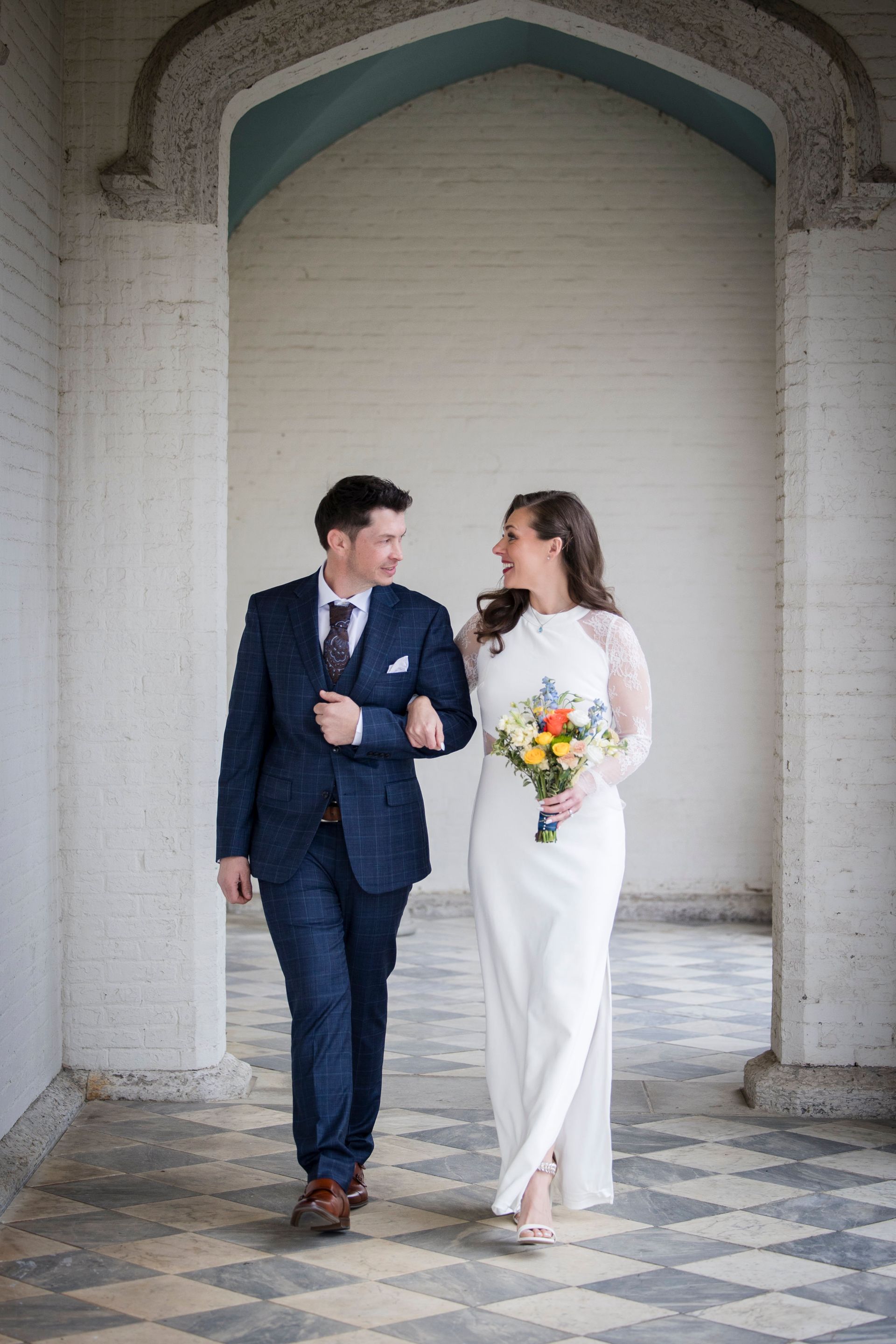 Couple in wedding attire walking hand in hand under an archway, smiling at each other.