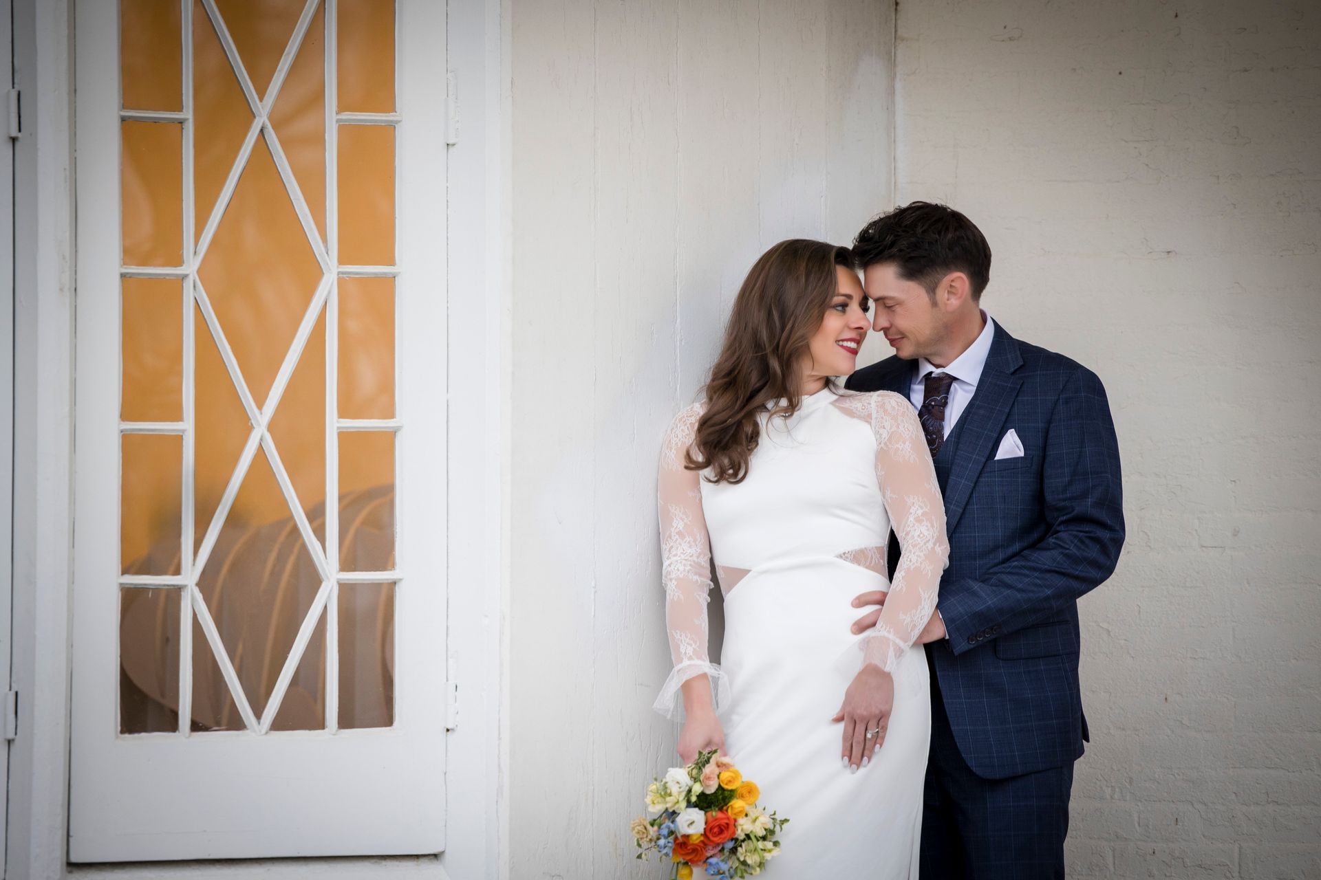 Bride and groom posing by a white doorway, smiling close together, bride holding a colorful bouquet