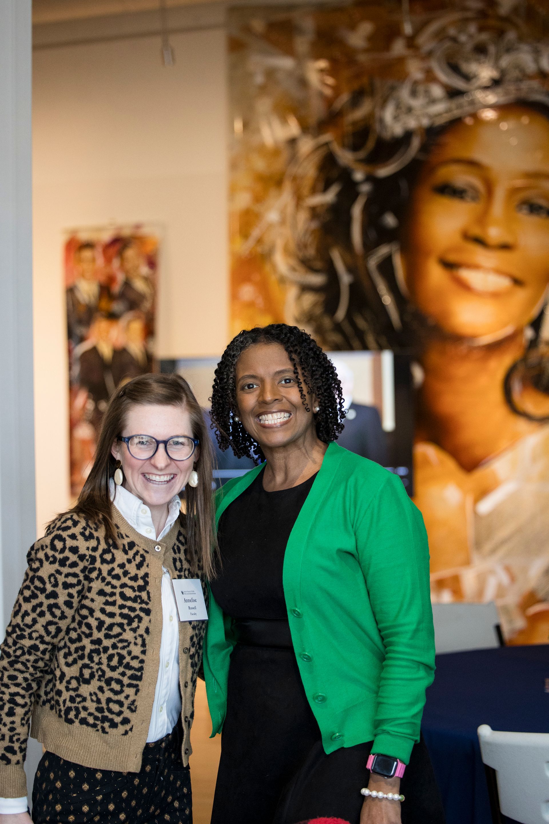 Two women smiling and posing indoors in front of a large wall portrait.