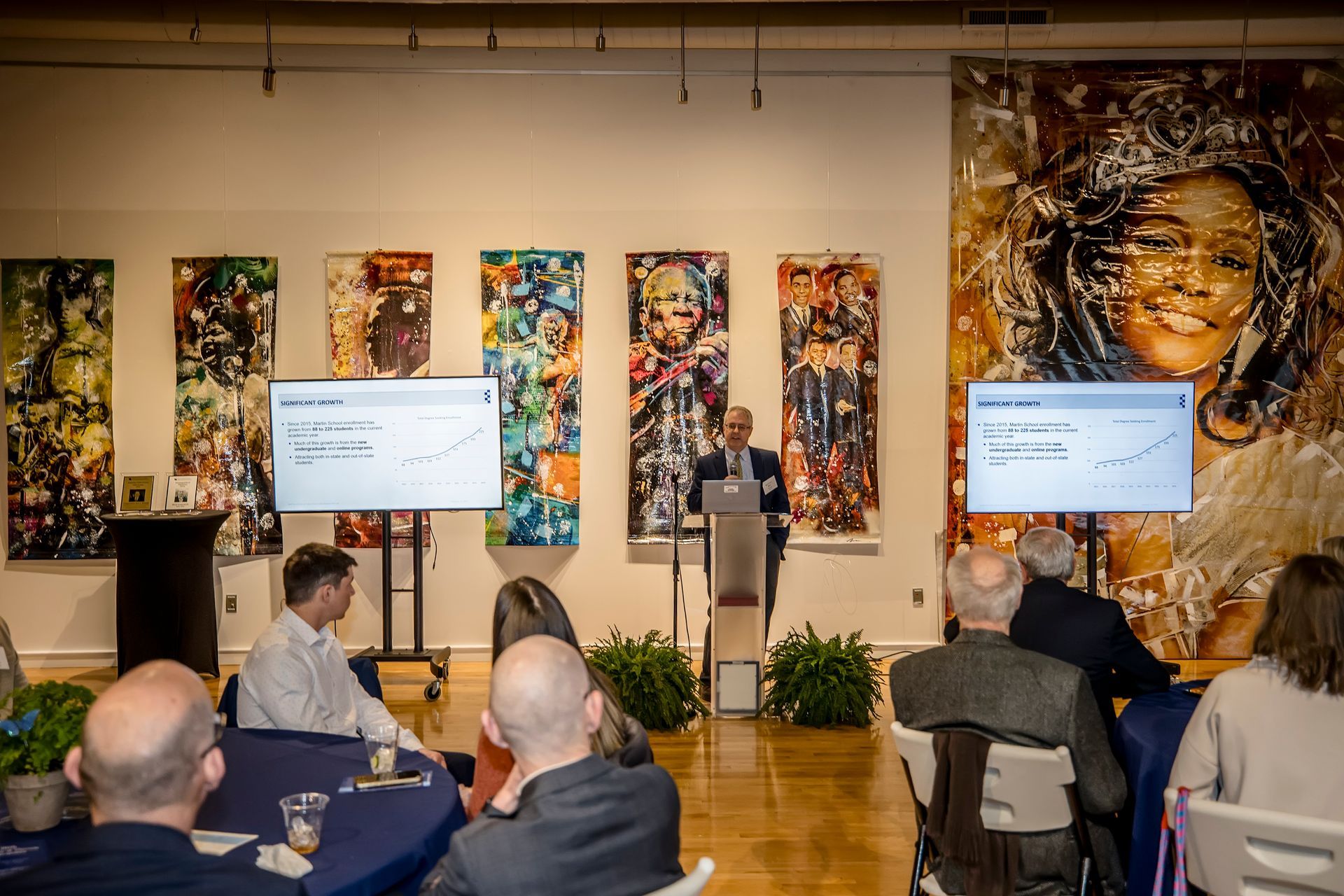 Speaker at podium in gallery space with colorful artwork and seated audience watching presentation