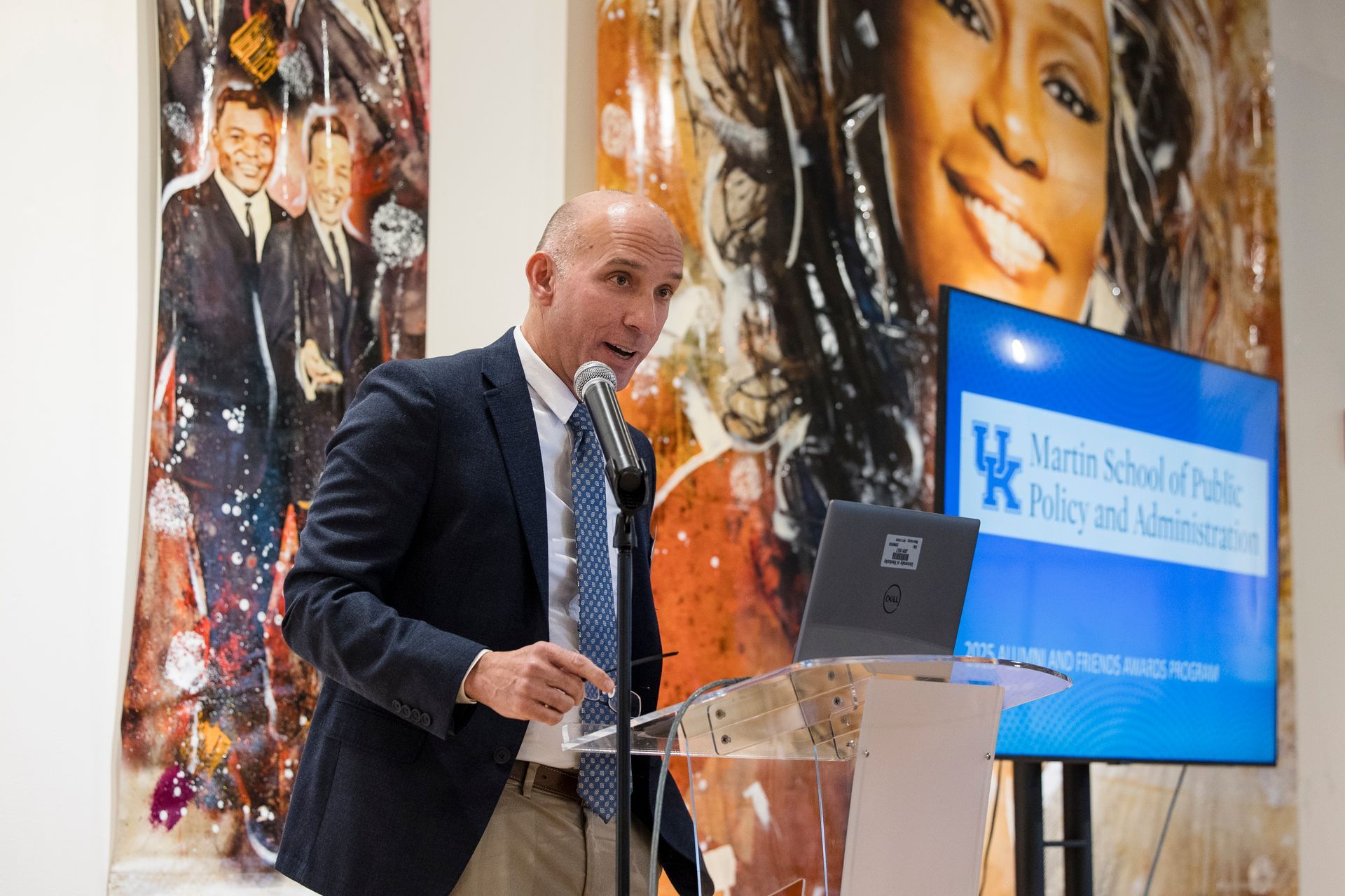 Speaker at a podium giving a presentation beside a blue display screen in a conference setting