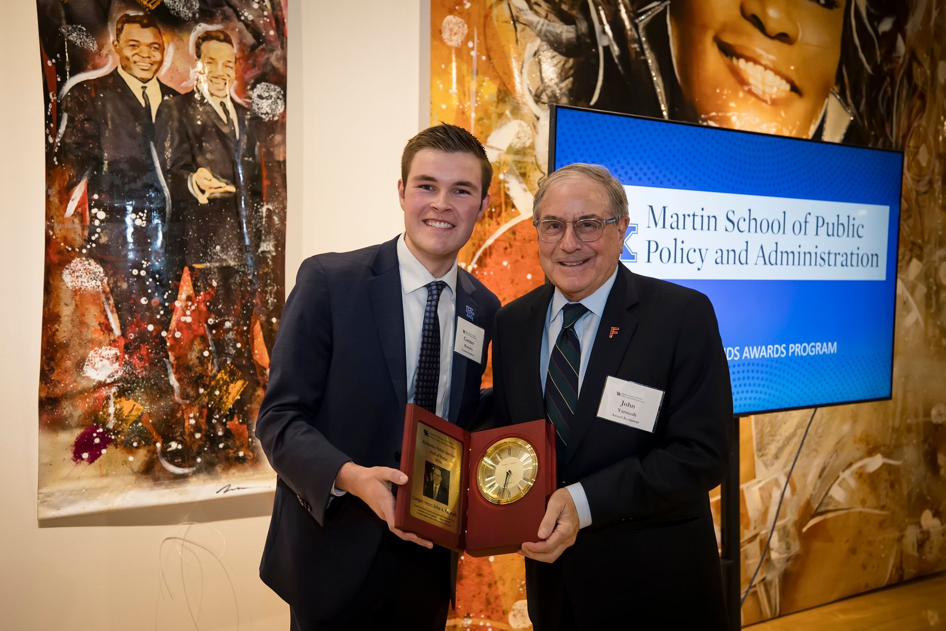 Two men in suits hold a plaque at an awards event, with a blue Marian School of Business and Administration sign behind them.