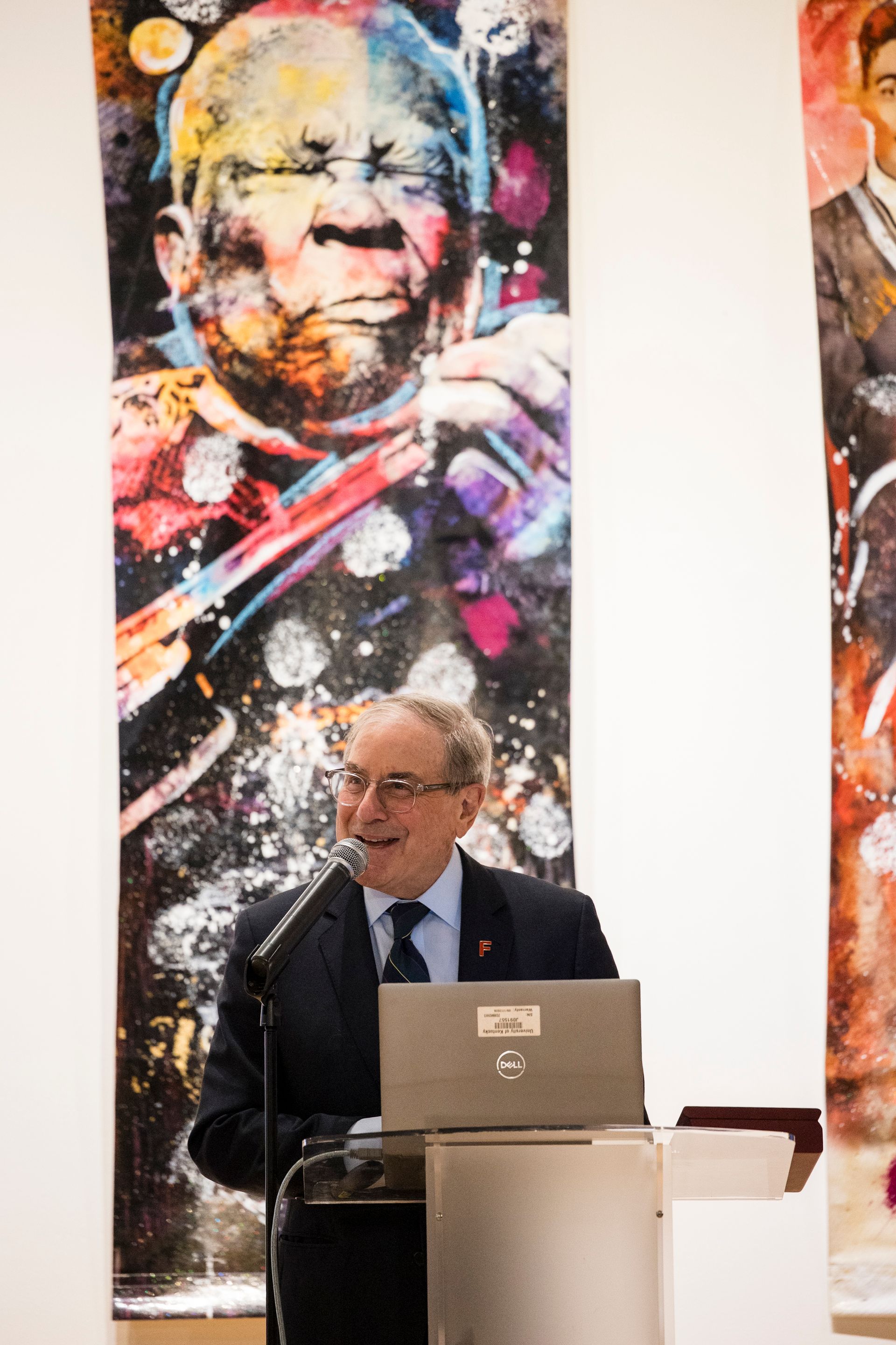 Man speaking at podium with laptop, framed by colorful portrait artwork on a white wall.