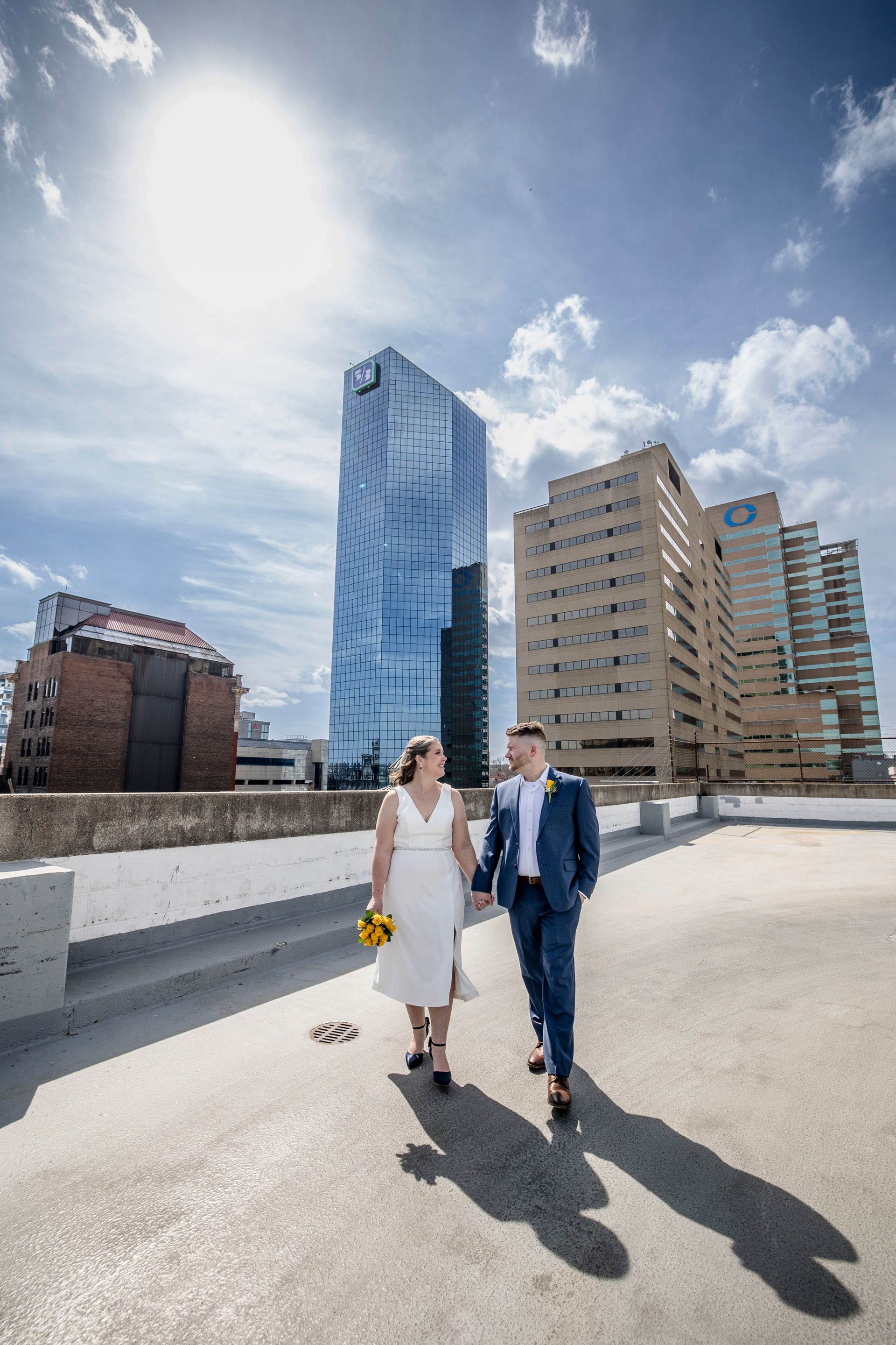 Couple in wedding attire walking on a rooftop with a city skyline and tall glass building behind them