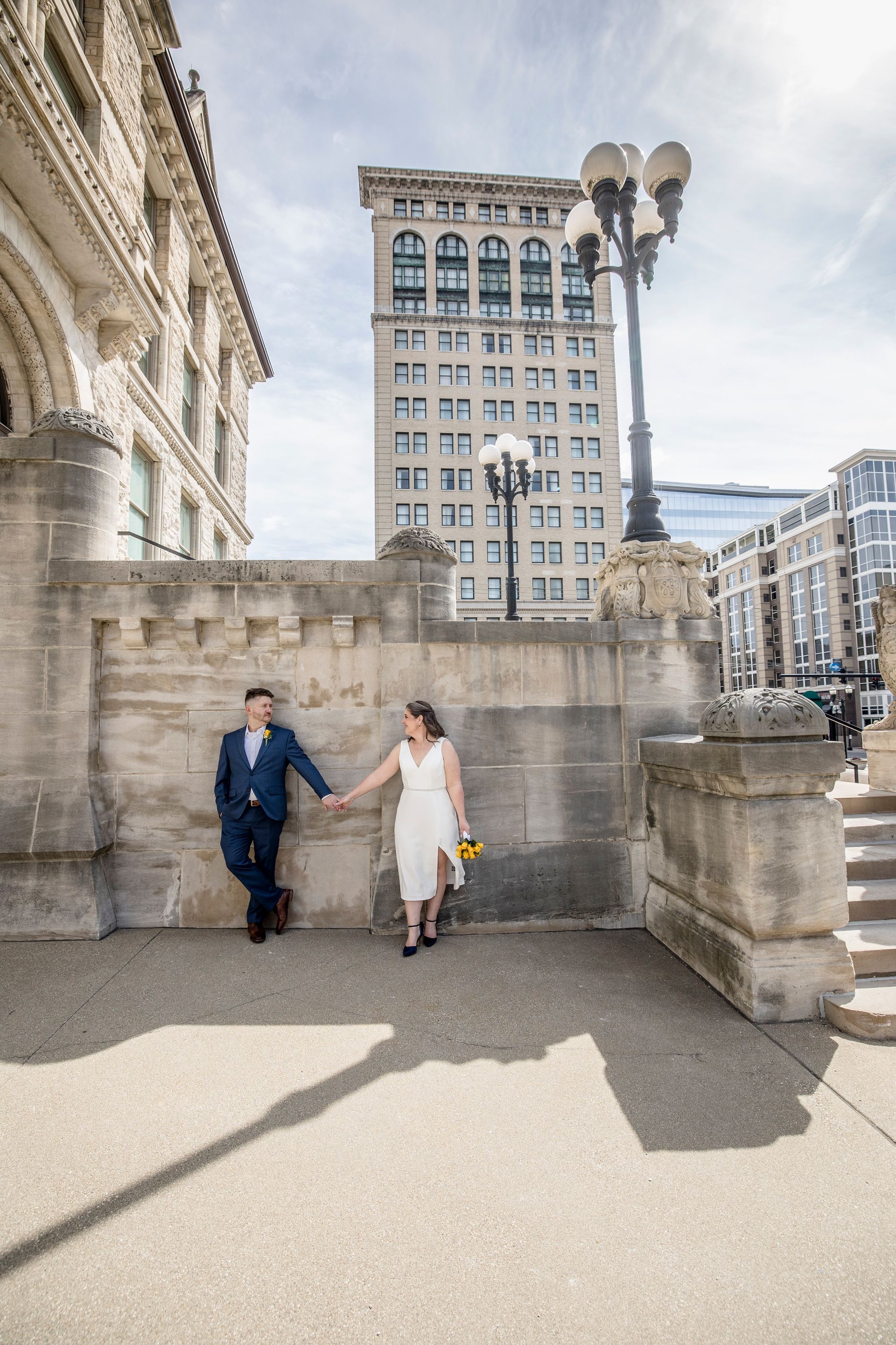 Couple holding hands walking beside stone buildings in a sunlit city plaza