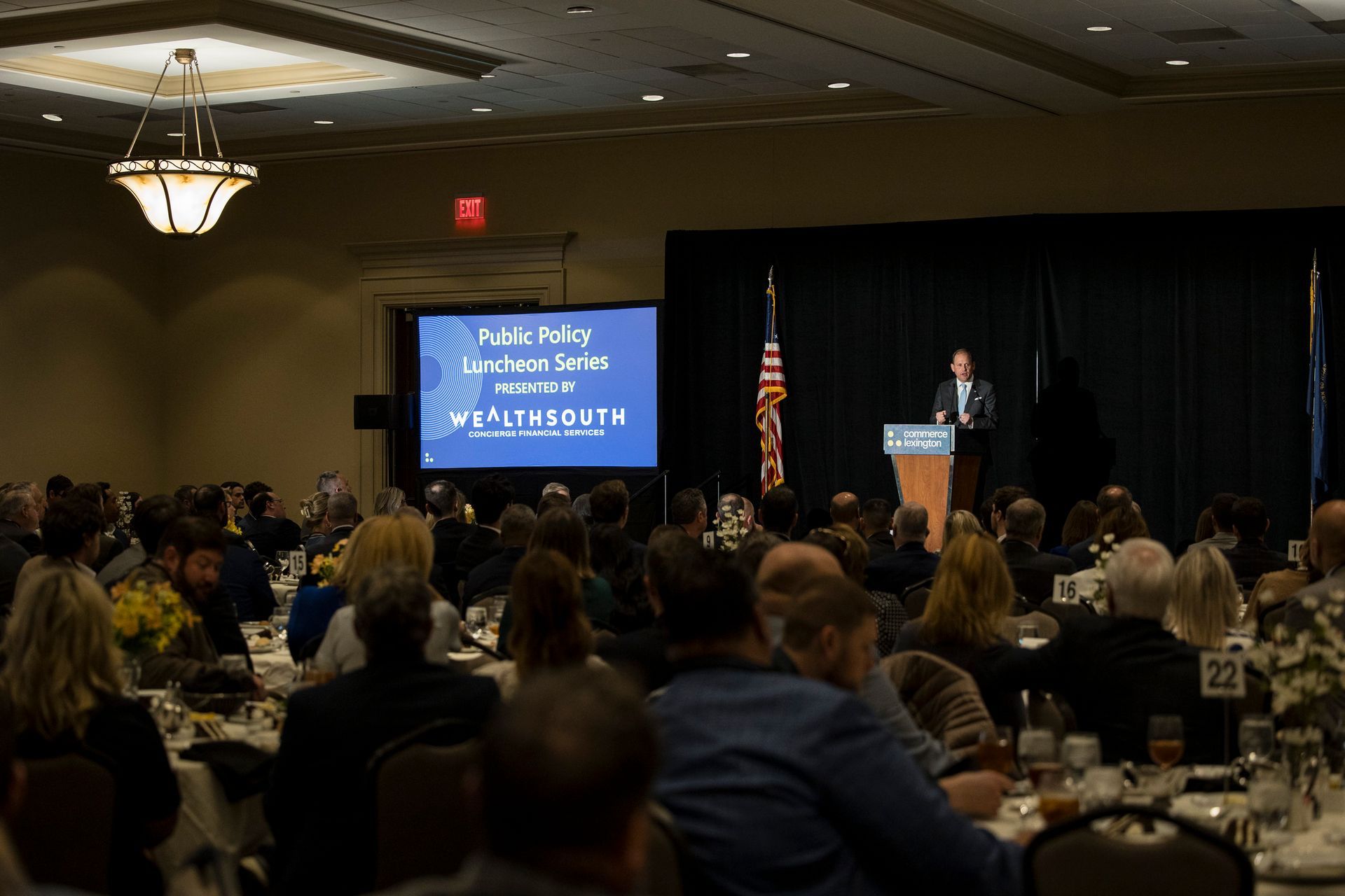 Conference audience watching a speaker at a podium beside a blue presentation screen in a ballroom