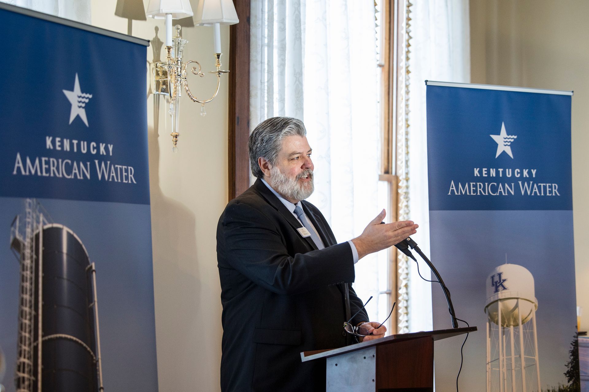 Man speaking at a podium at a Kentucky American Water event, flanked by blue company banners.