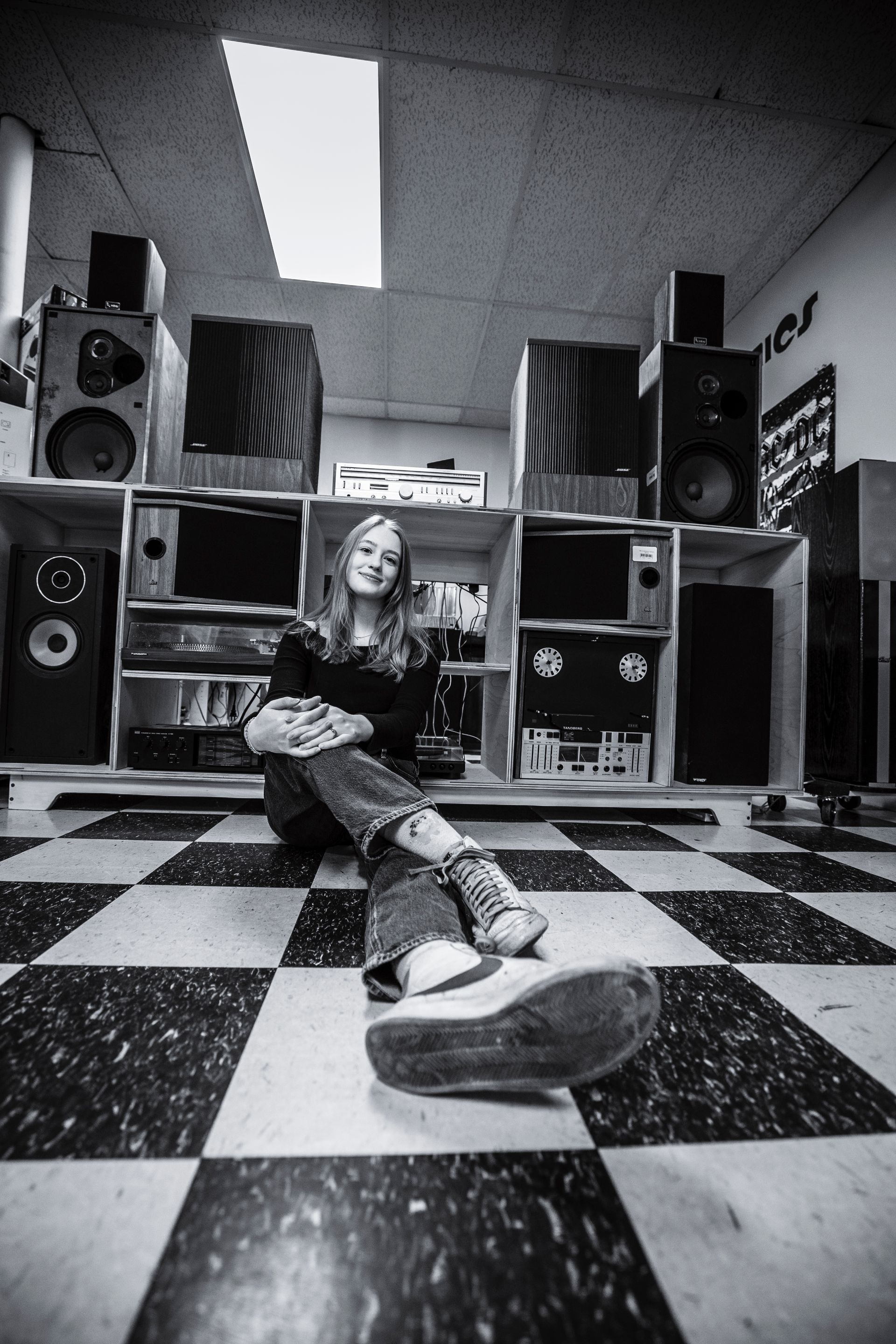 Person reclining on a checkered floor in a room lined with speakers and shelves.
