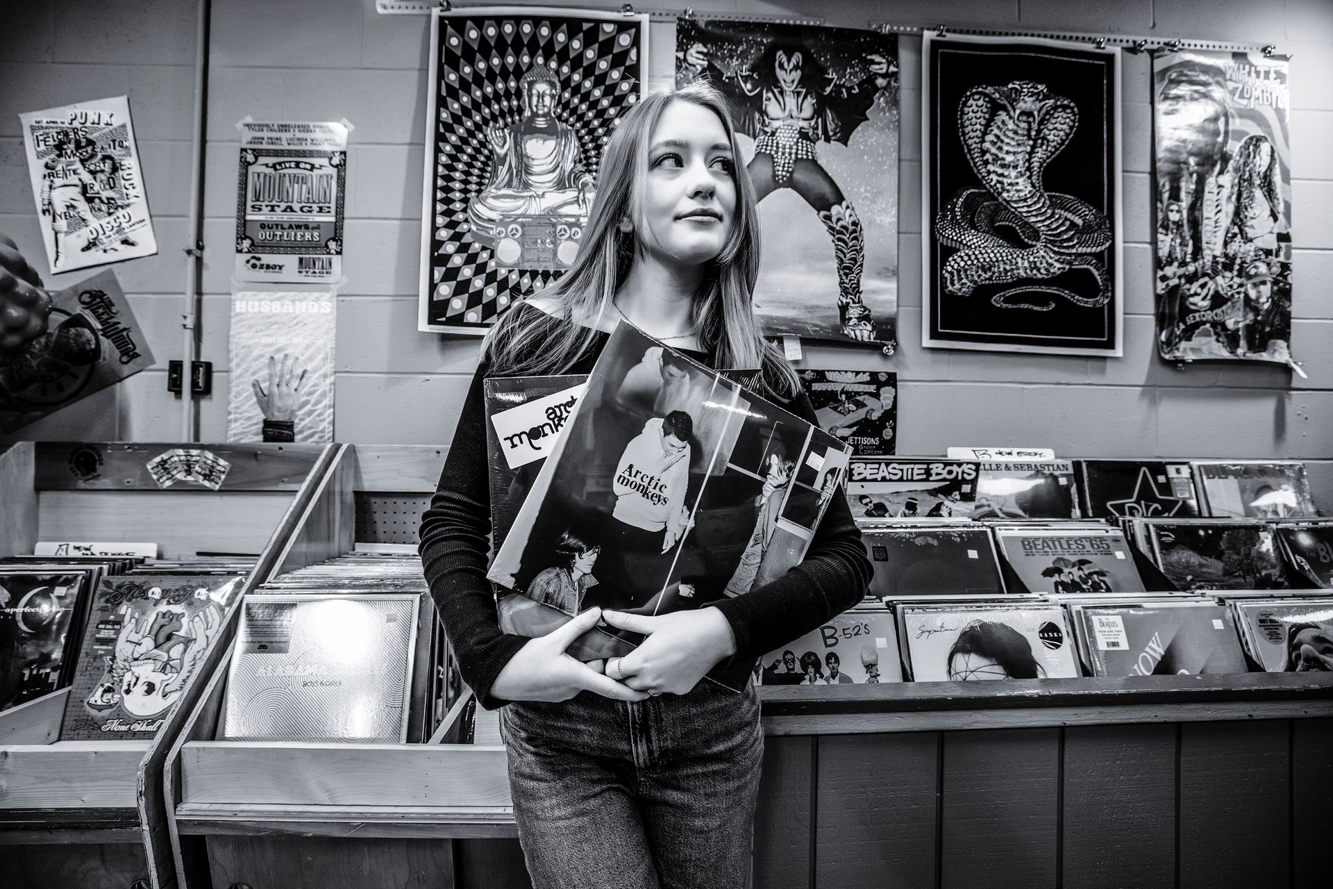Woman holding a skateboard in a record store, standing beside bins of vinyl albums and wall posters.