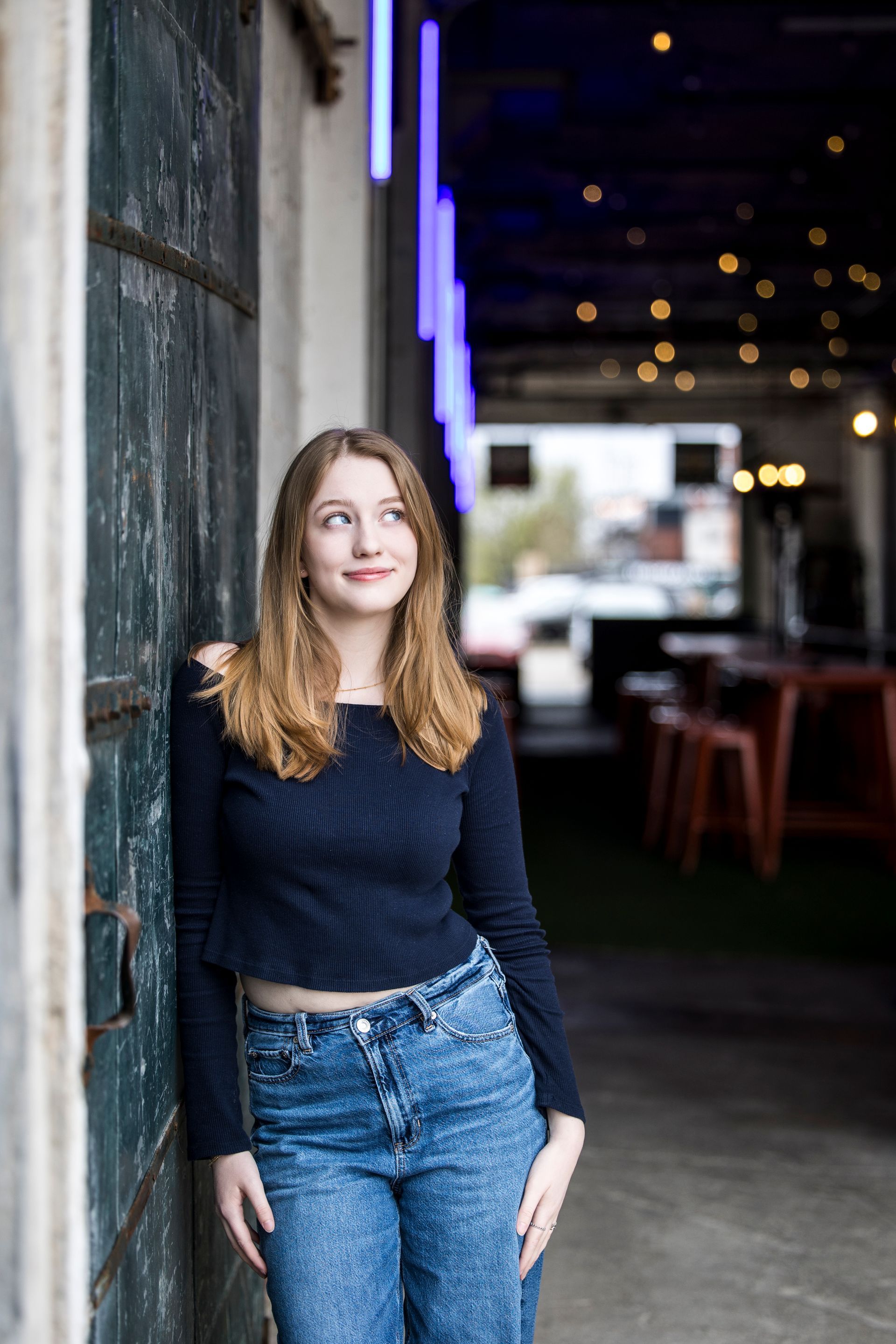 Young woman in a navy sweater and jeans leaning against a wall in a dimly lit hallway