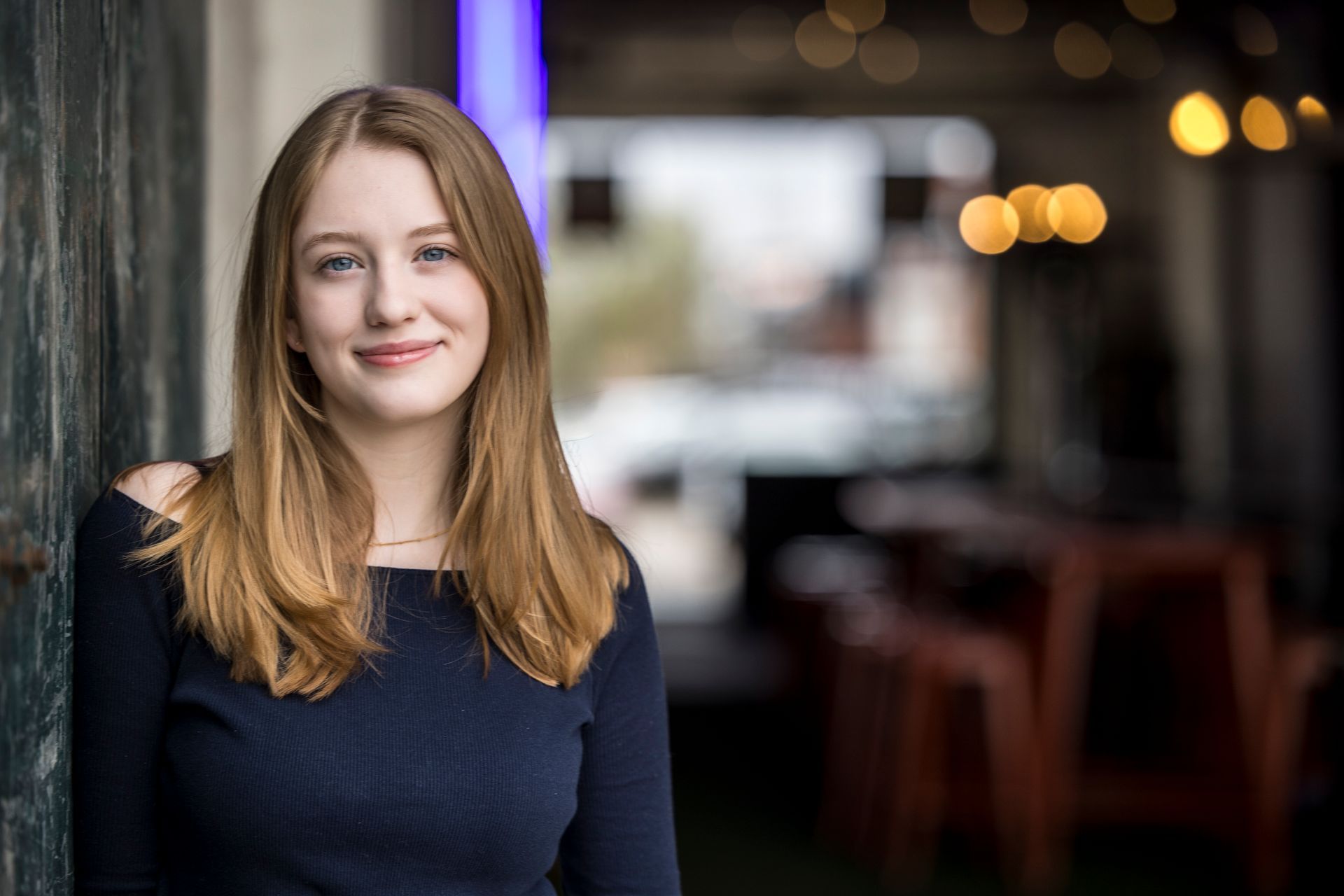 Woman with long red hair smiling in a dimly lit cafe, wearing a dark blue top.