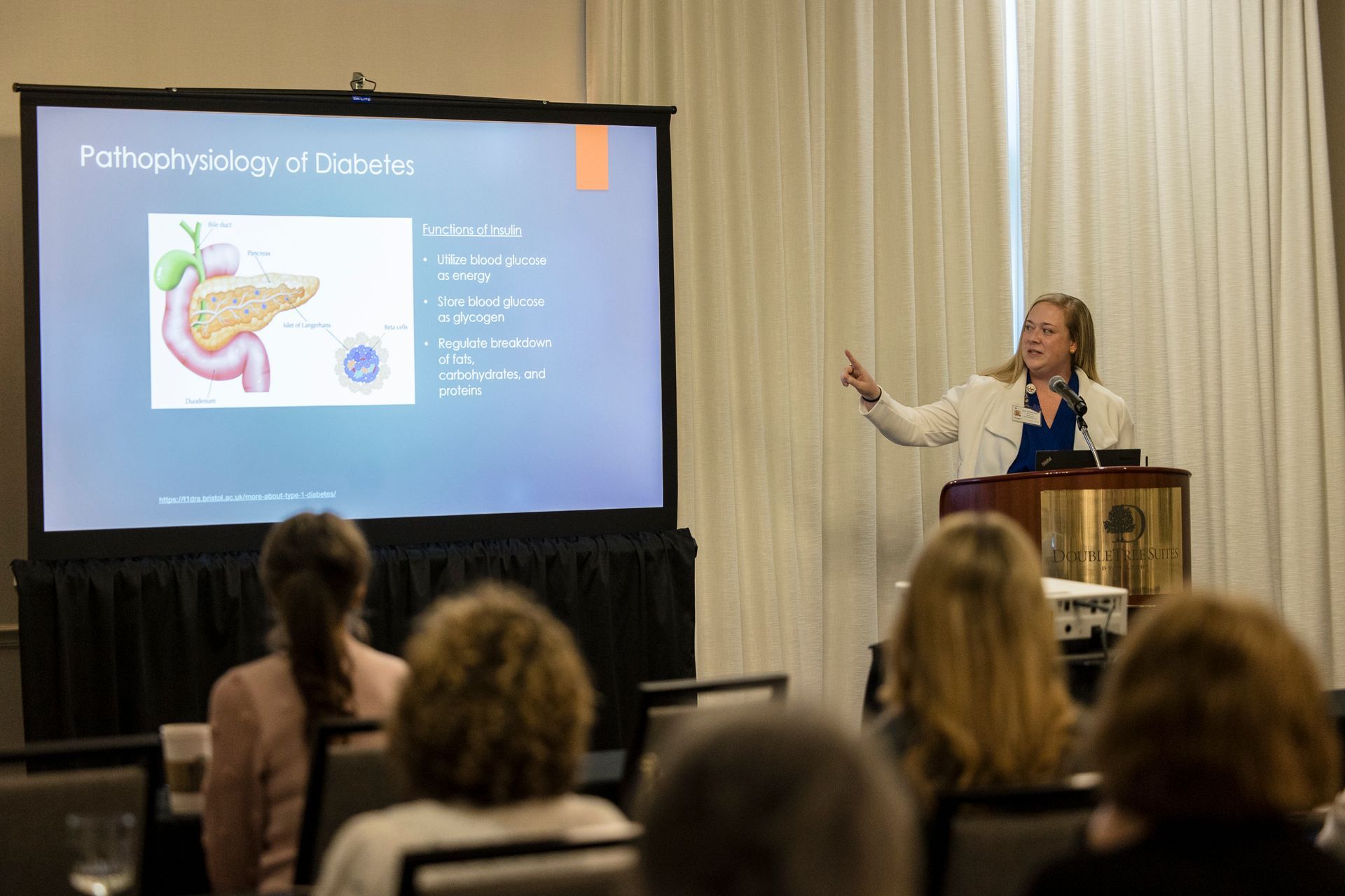 Speaker presenting at a conference, gesturing beside a slide titled “Pathophysiology of Diabetes”