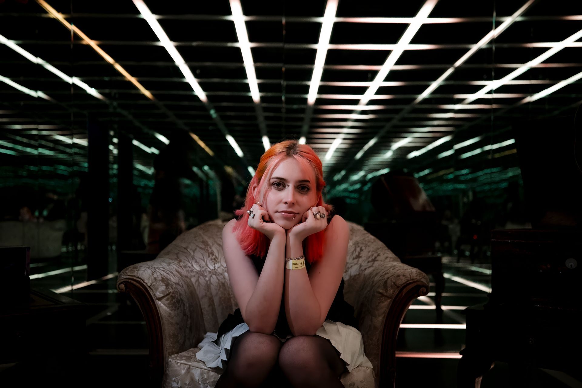 Young woman seated on a couch in a dark neon-lit room, resting her chin on her hands, looking at the camera