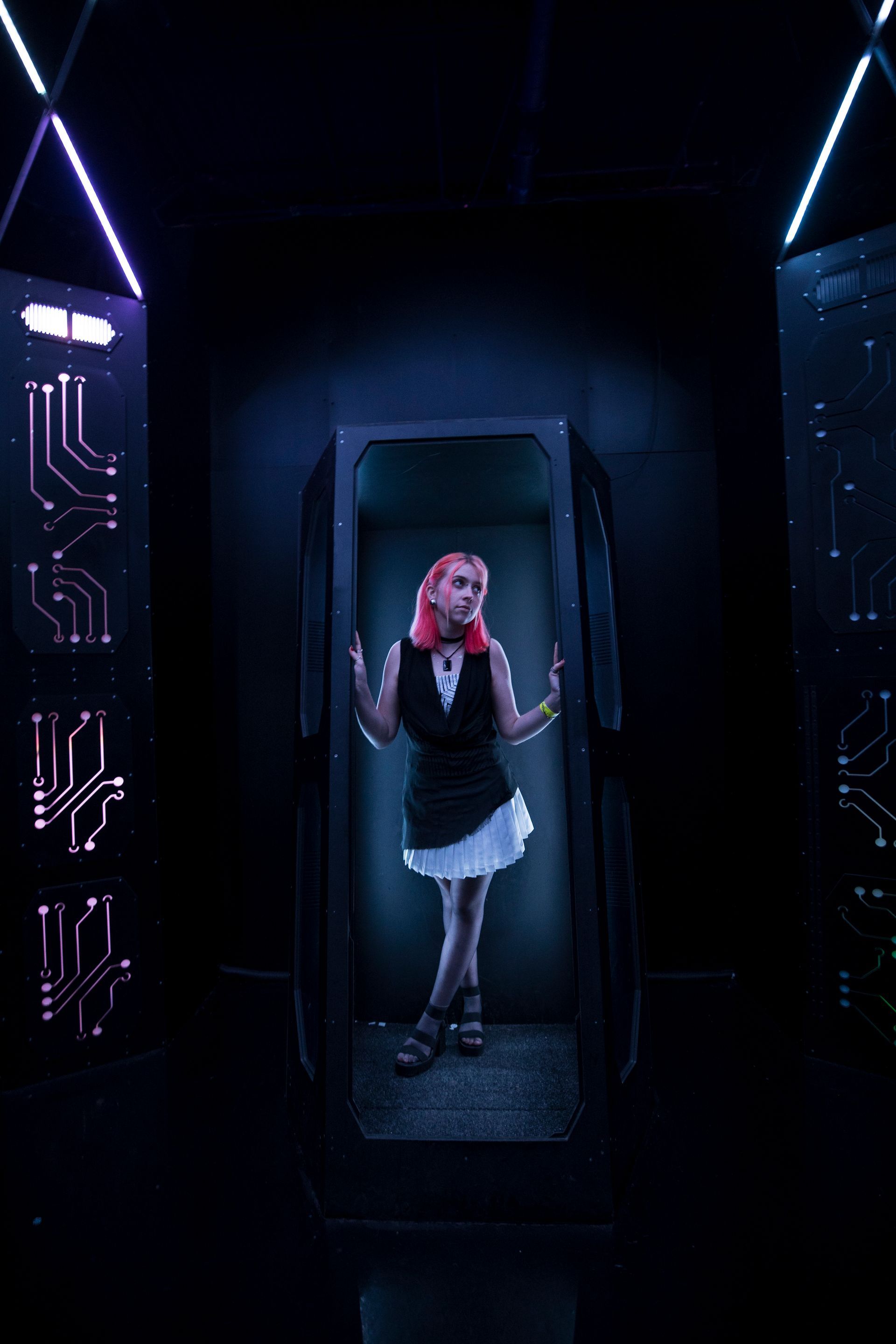 Woman stands in a dark futuristic doorway with neon lights, holding one hand up and wearing a black dress.