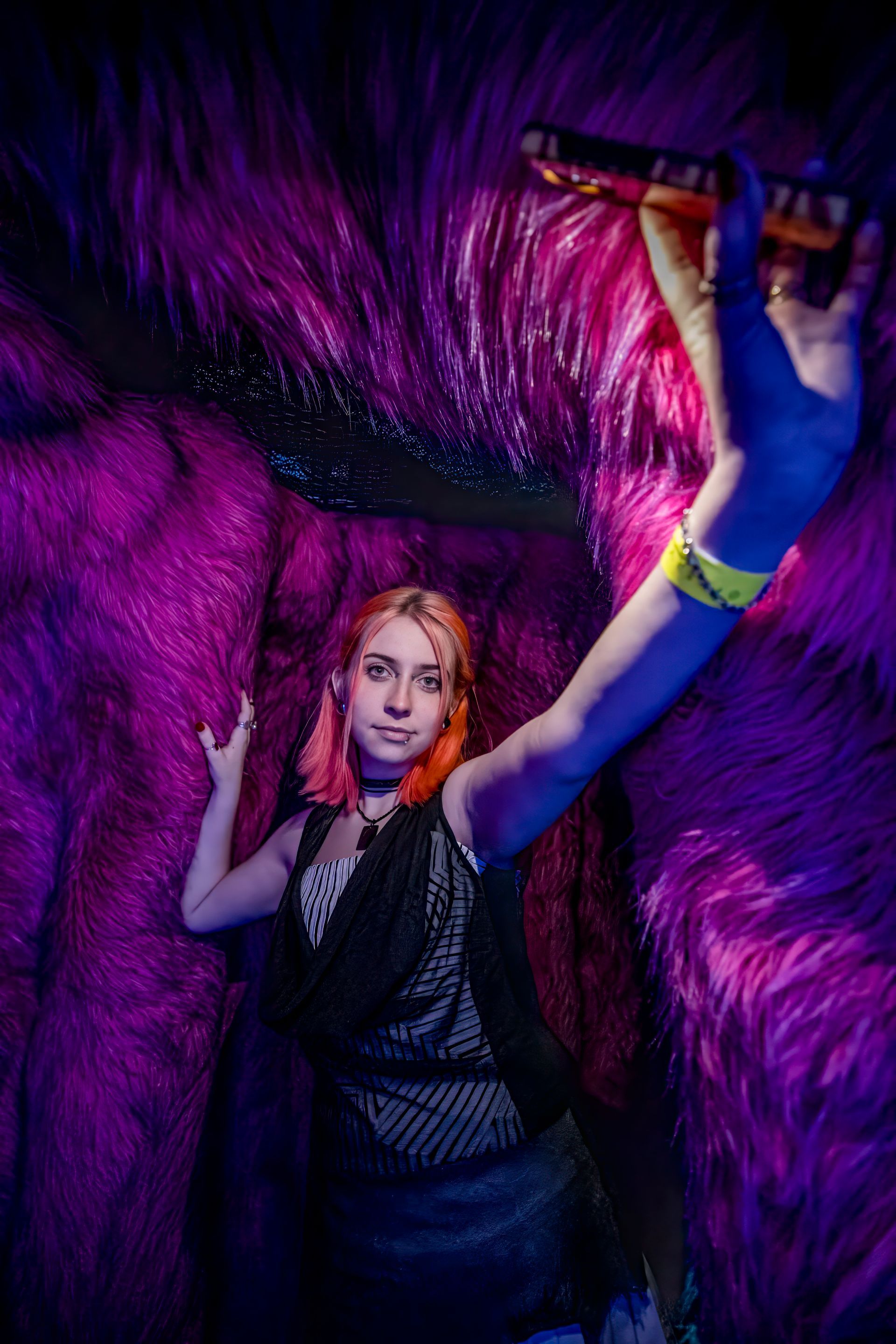 Person posing with raised arm against a vivid purple feathered backdrop, holding a flute-like object.