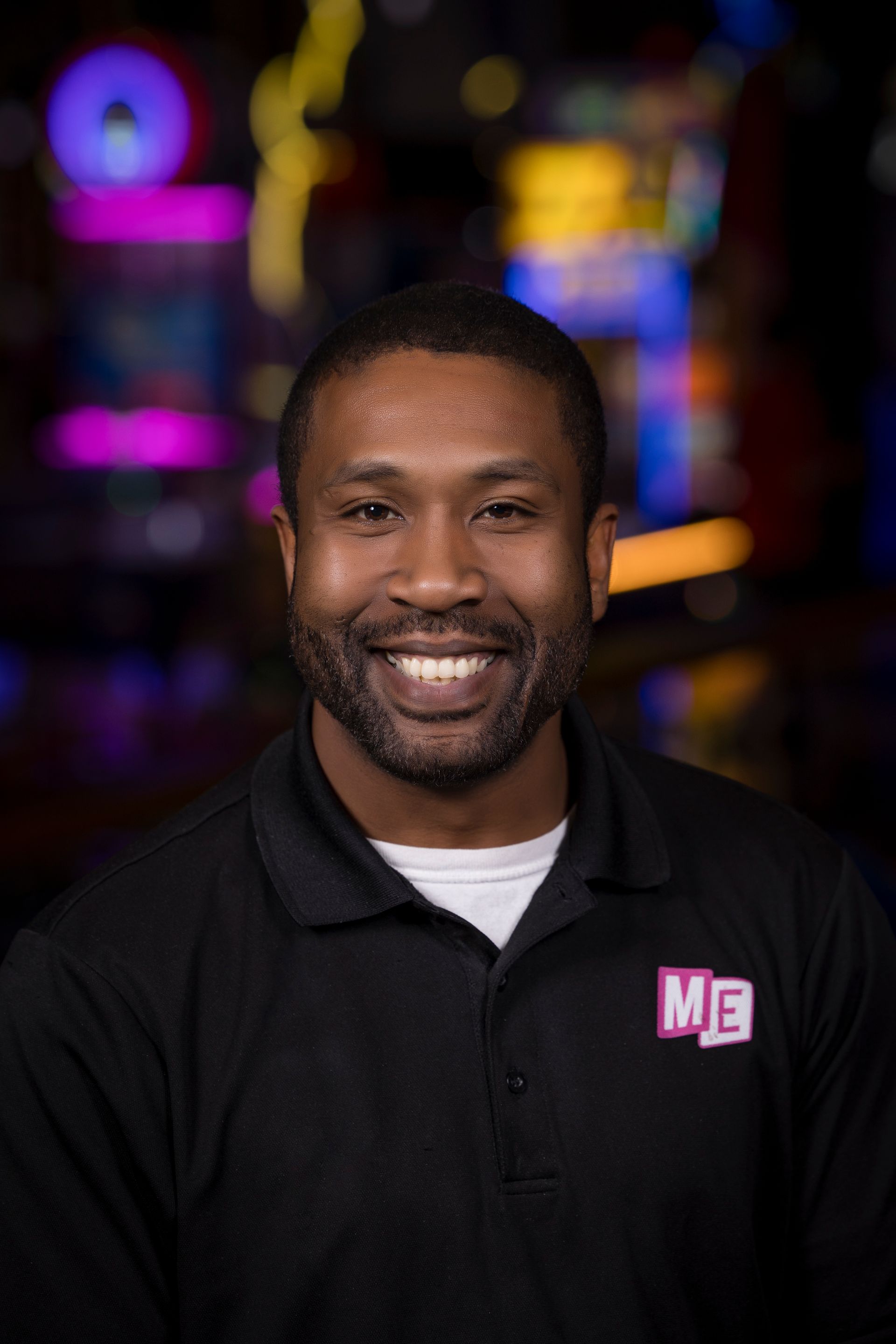 Smiling man in a black polo with an “ME” logo, standing against a colorful blurred background