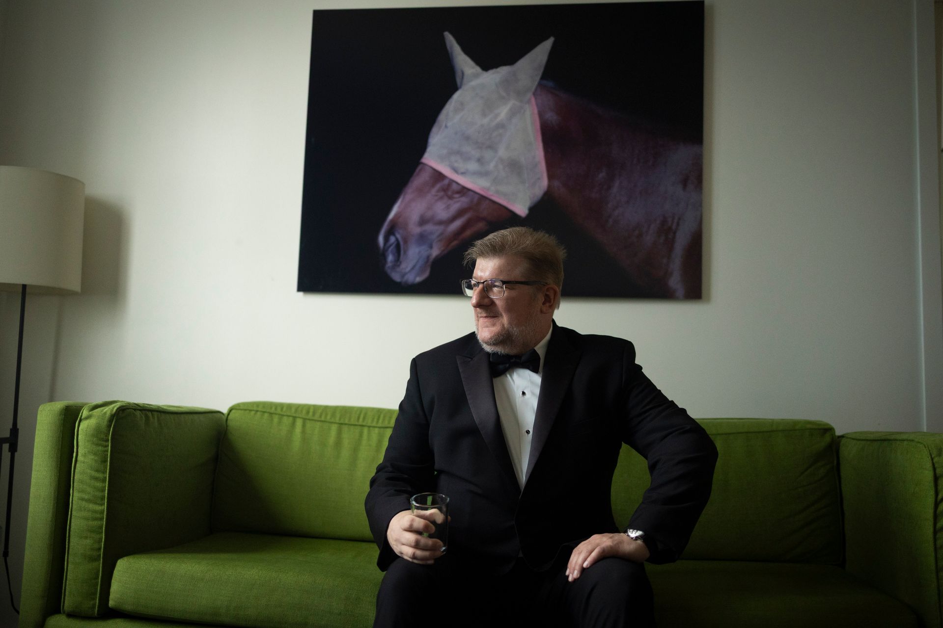 Man in a tuxedo sitting on a green sofa beneath a horse portrait.