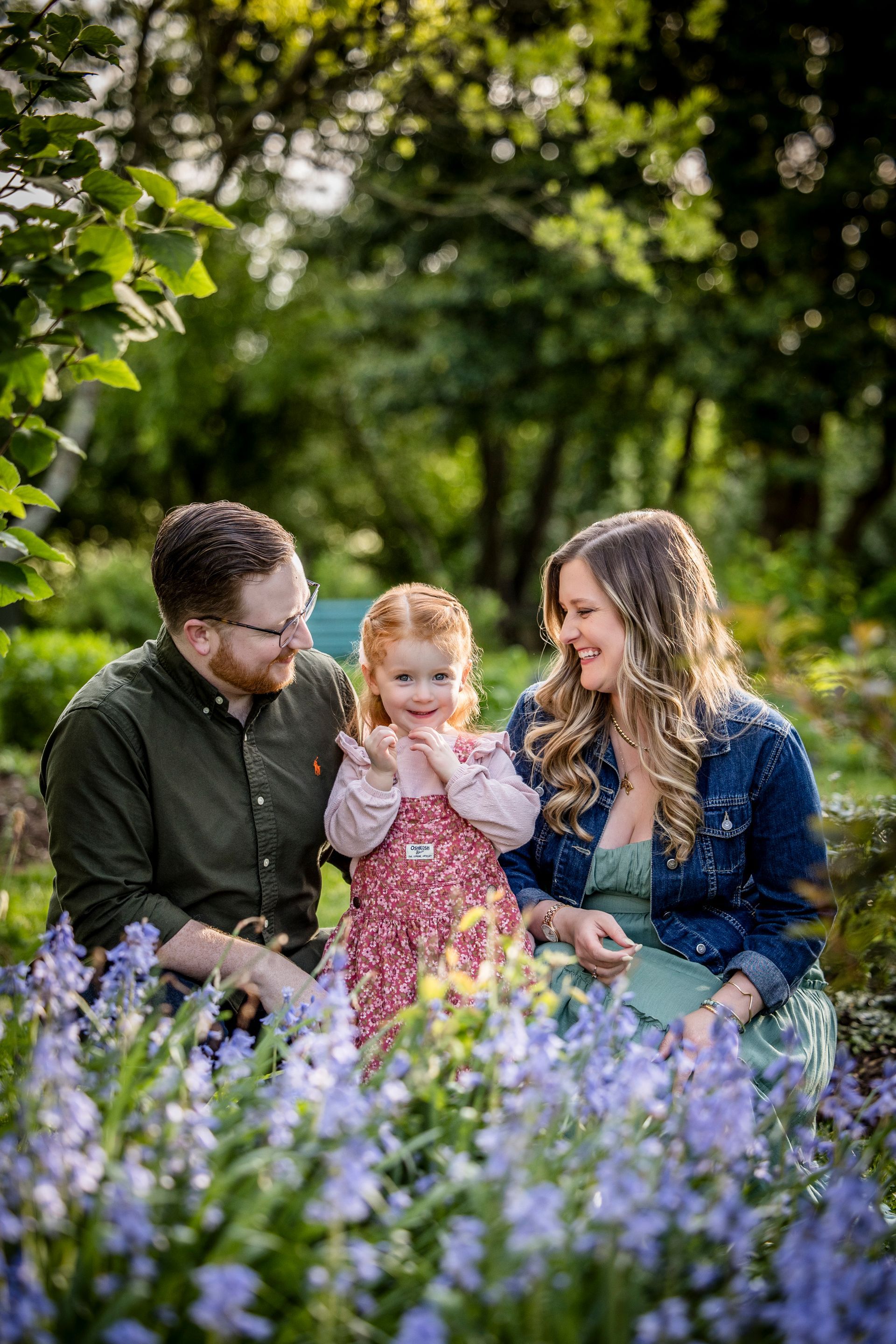 Family sitting in a flowered garden, smiling at a toddler among purple blooms.