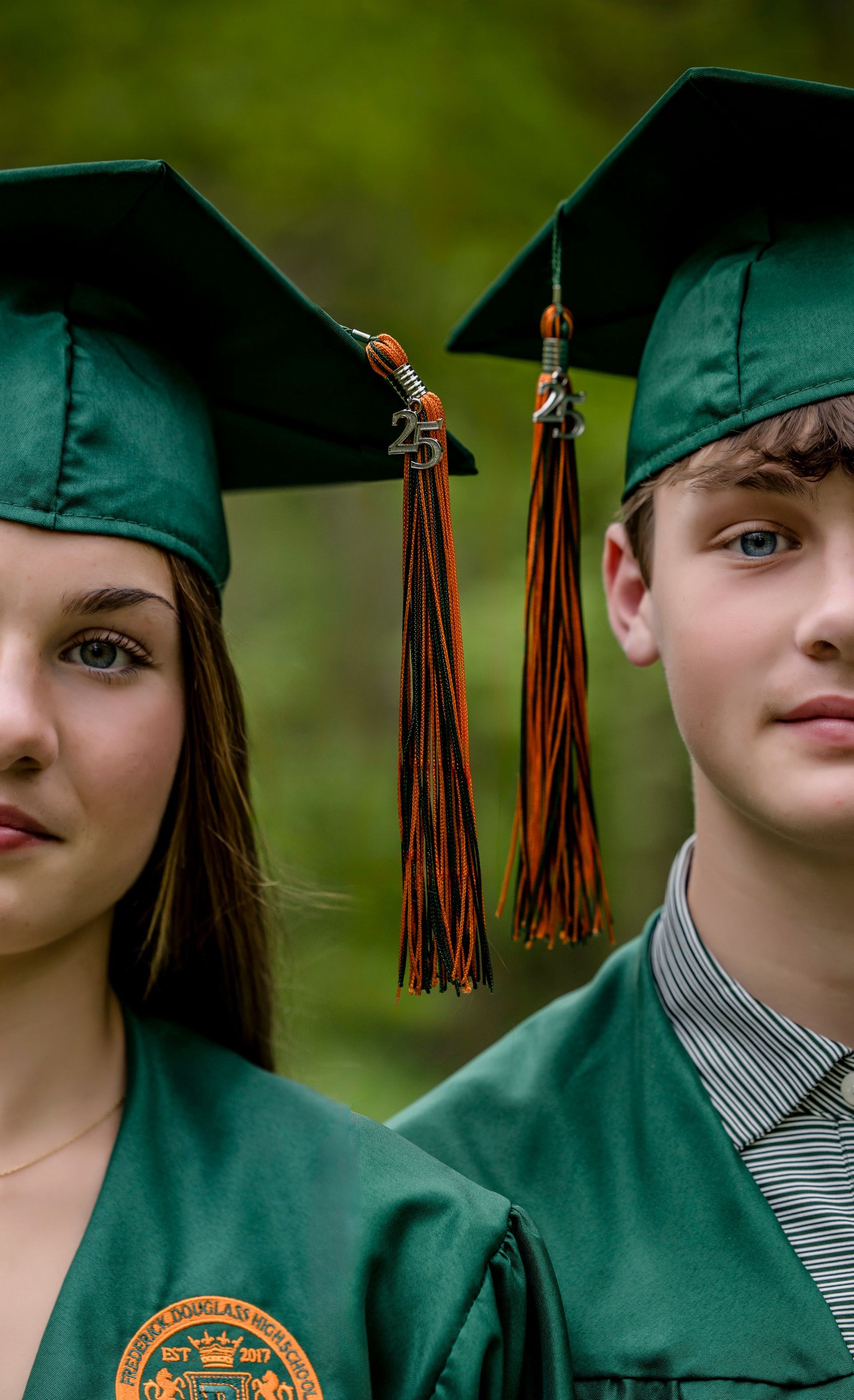 Two graduates in green caps and gowns posing side by side against a dark green background