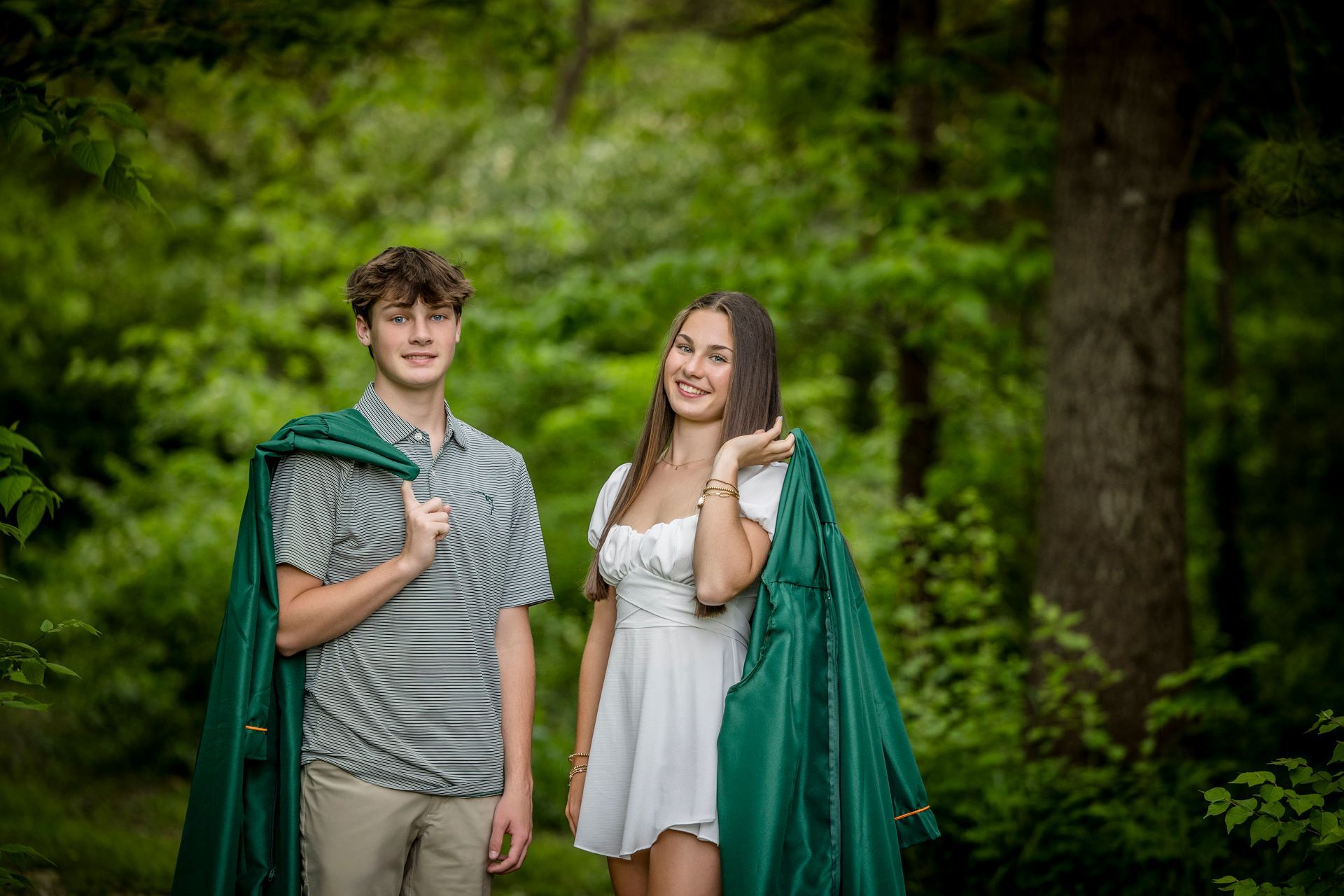 Two people in white outfits and green capes stand smiling in a lush forest