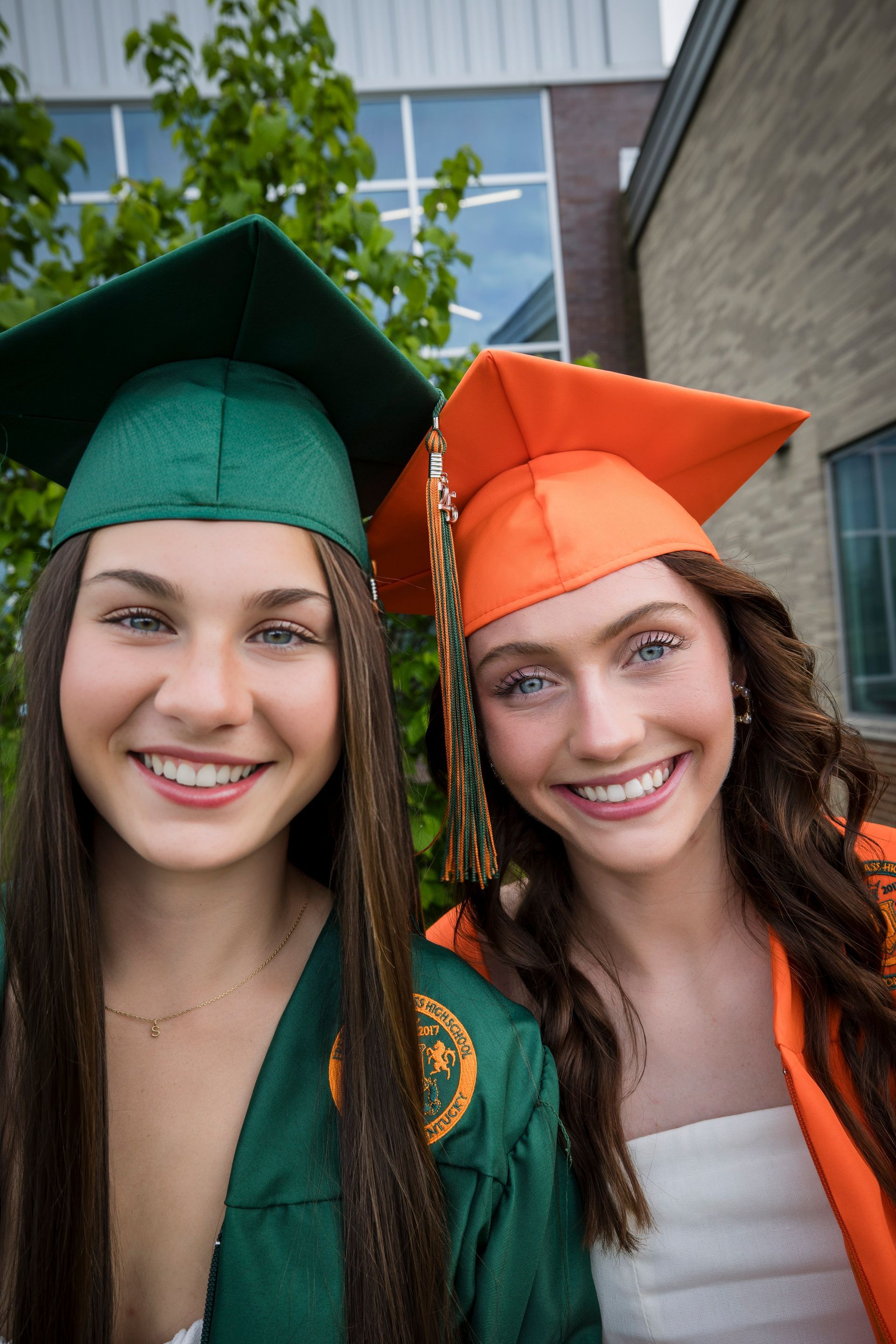 Two graduates in green and orange caps and gowns smiling outdoors
