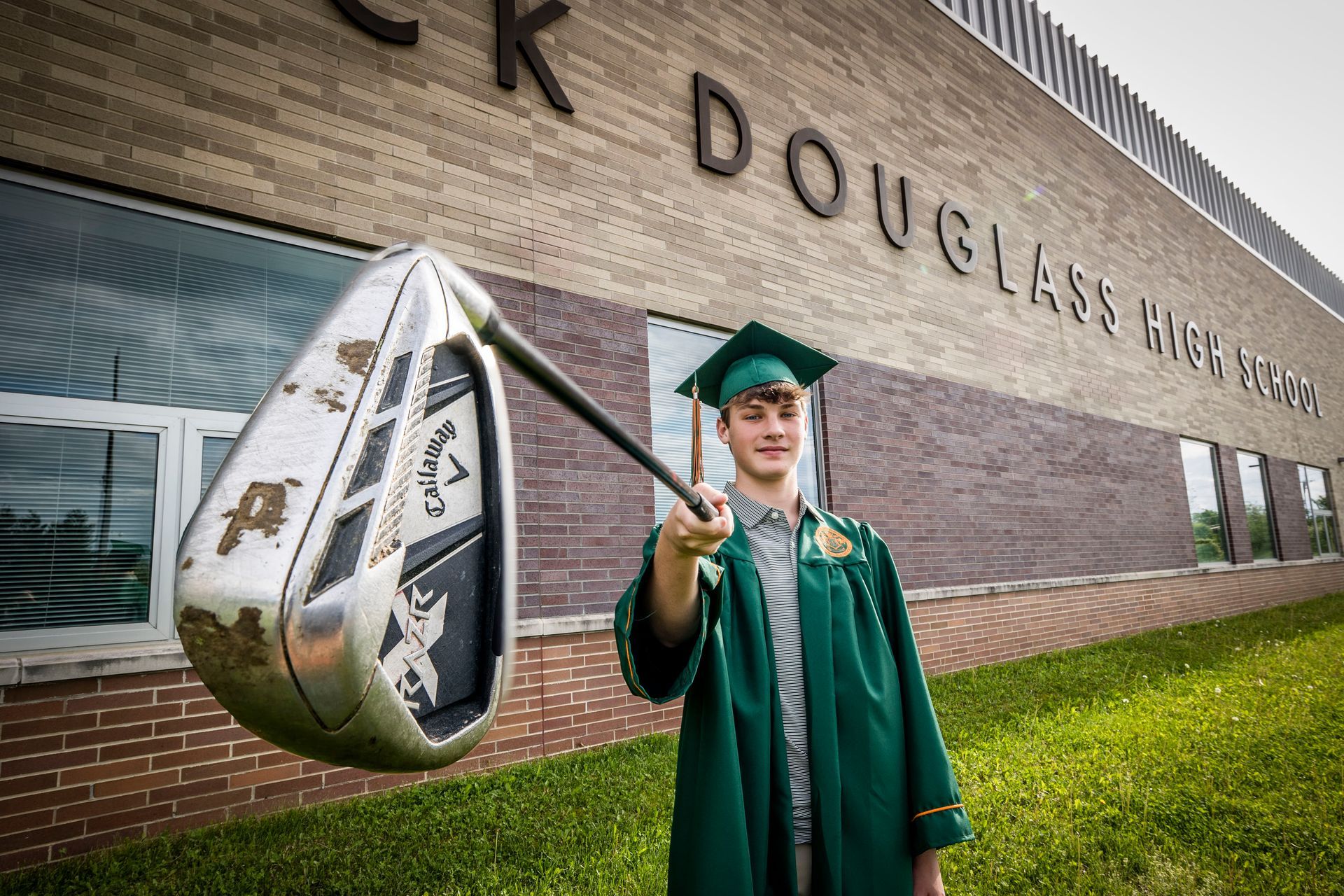 Graduate in green cap and gown holding a mace outside a brick building with large university lettering