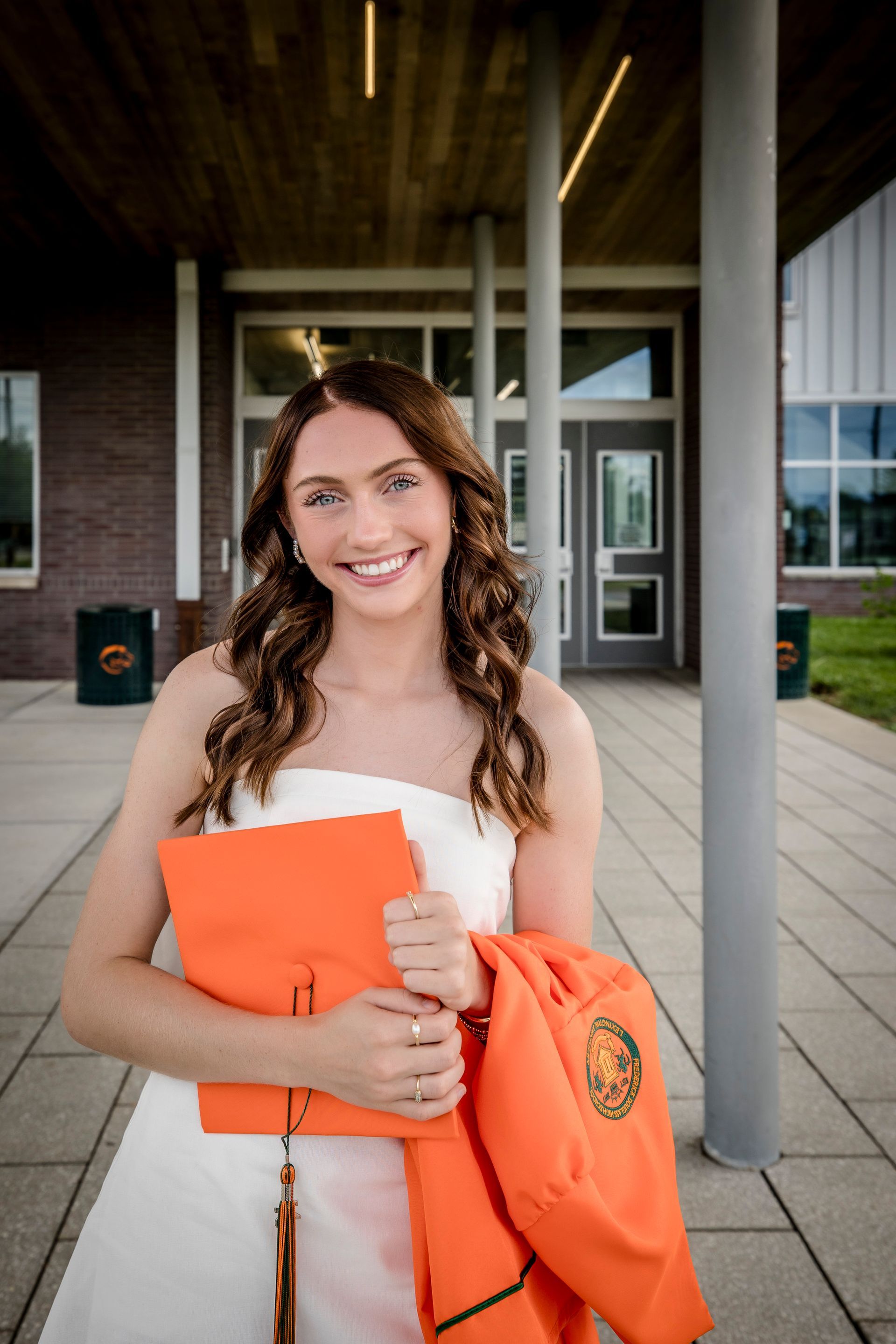 Smiling student holding an orange folder and sweatshirt outside a campus building
