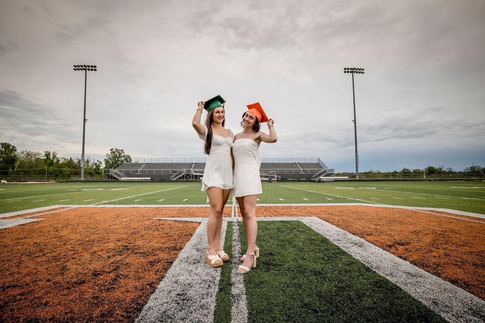 Two graduates in white dresses and caps stand on a football field, holding their mortarboards up