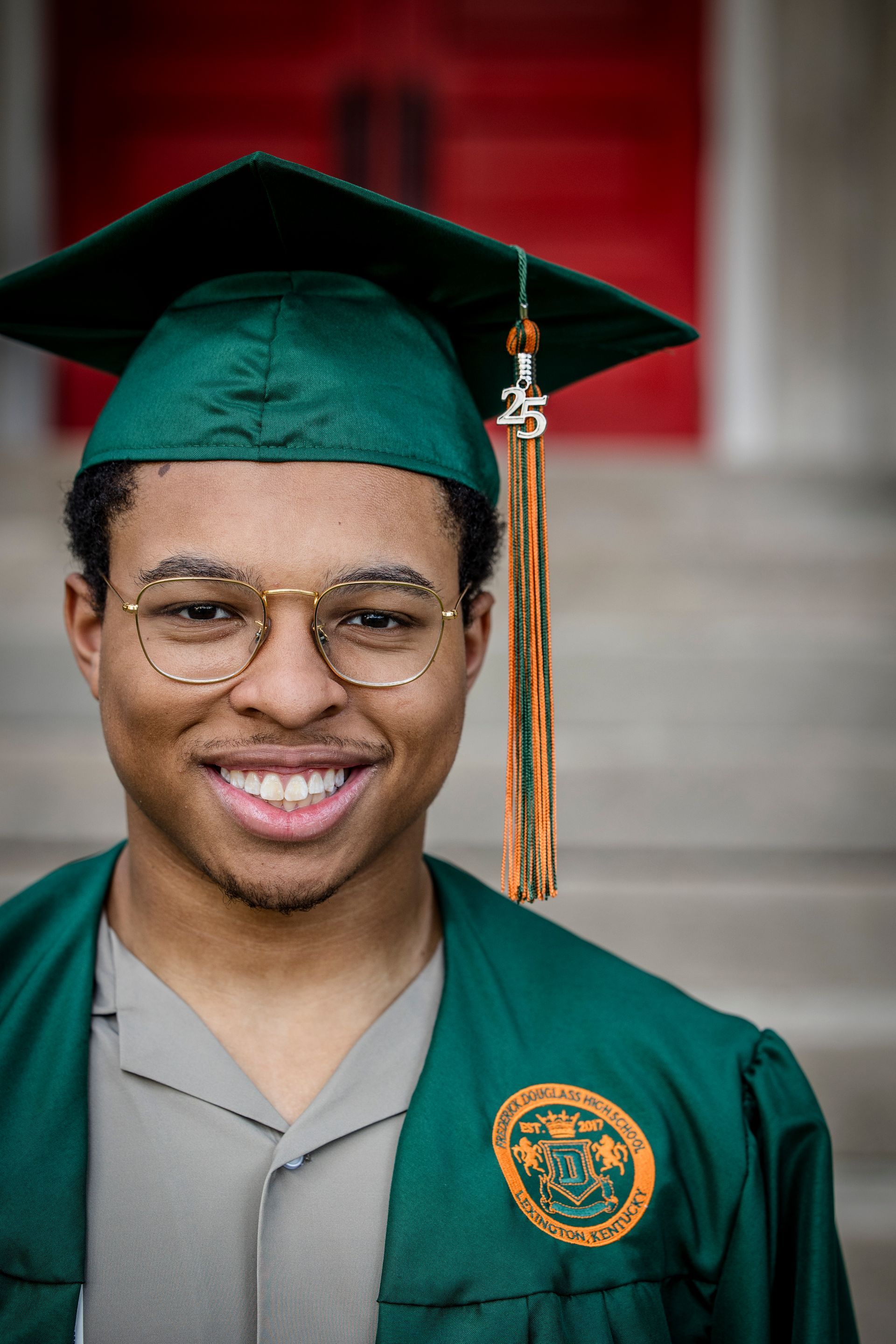 Portrait of a smiling graduate in a green cap and gown with a school emblem, standing on steps