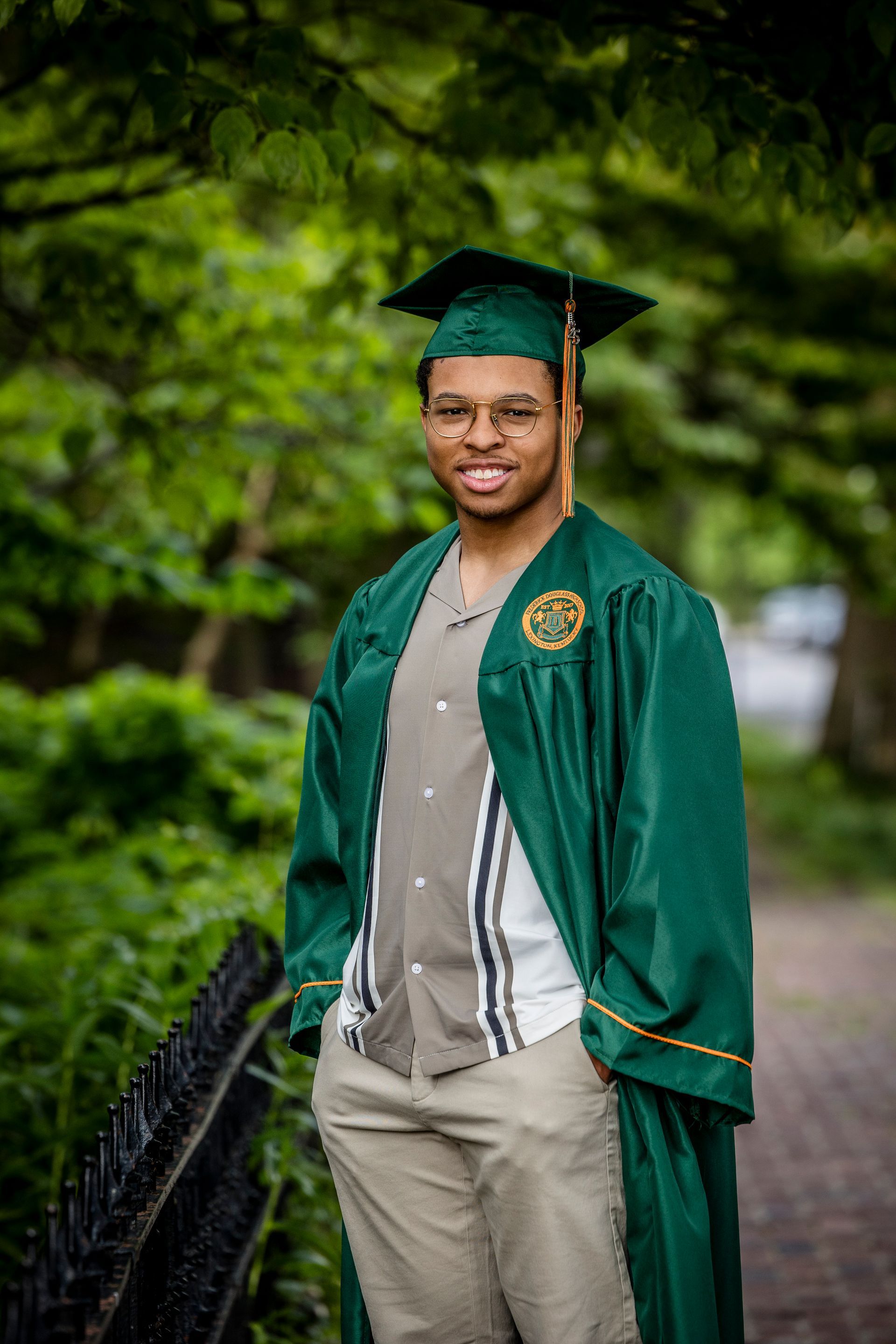 Graduate in green cap and gown standing outdoors on a bridge, smiling